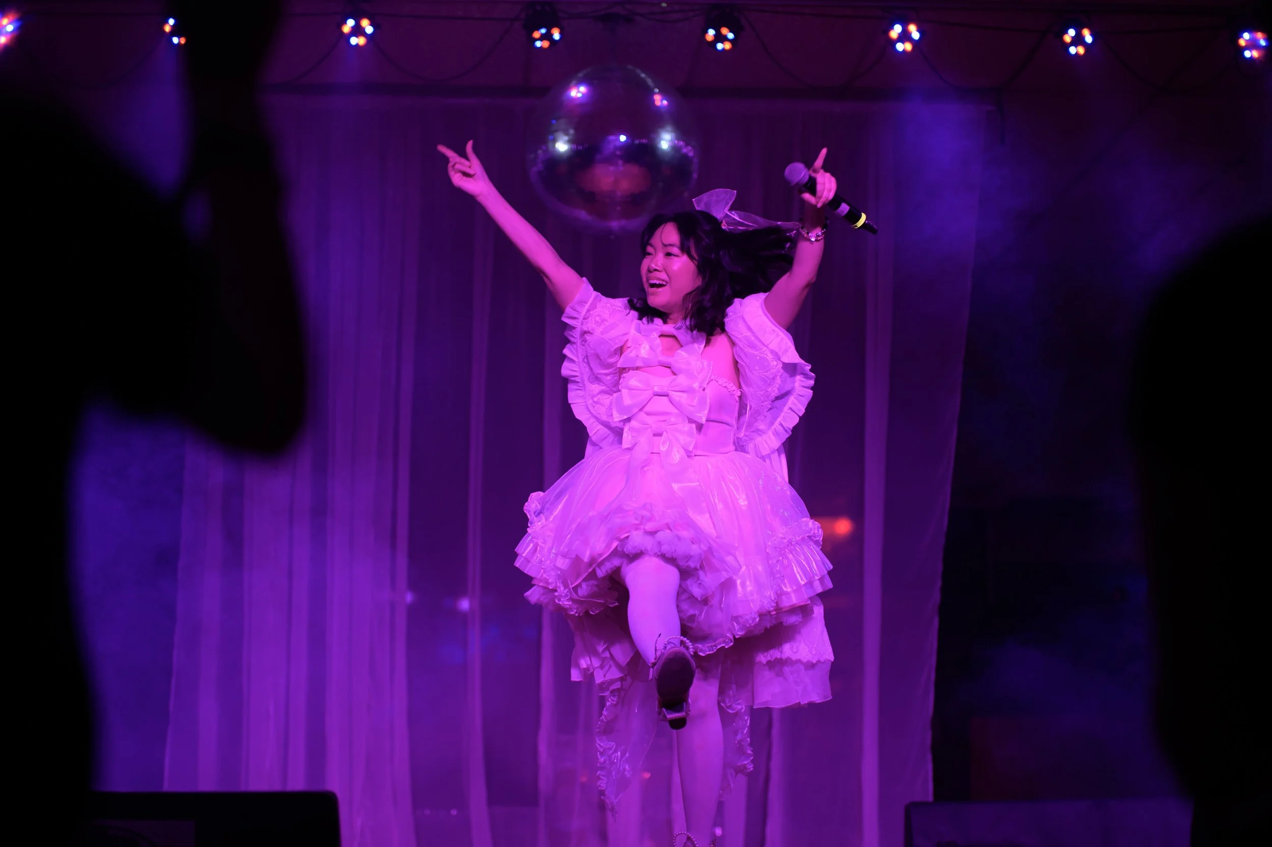 A woman in a frilly pink dress dancing and singing on stage with purple lighting, holding a microphone, and surrounded by string lights and a disco ball.