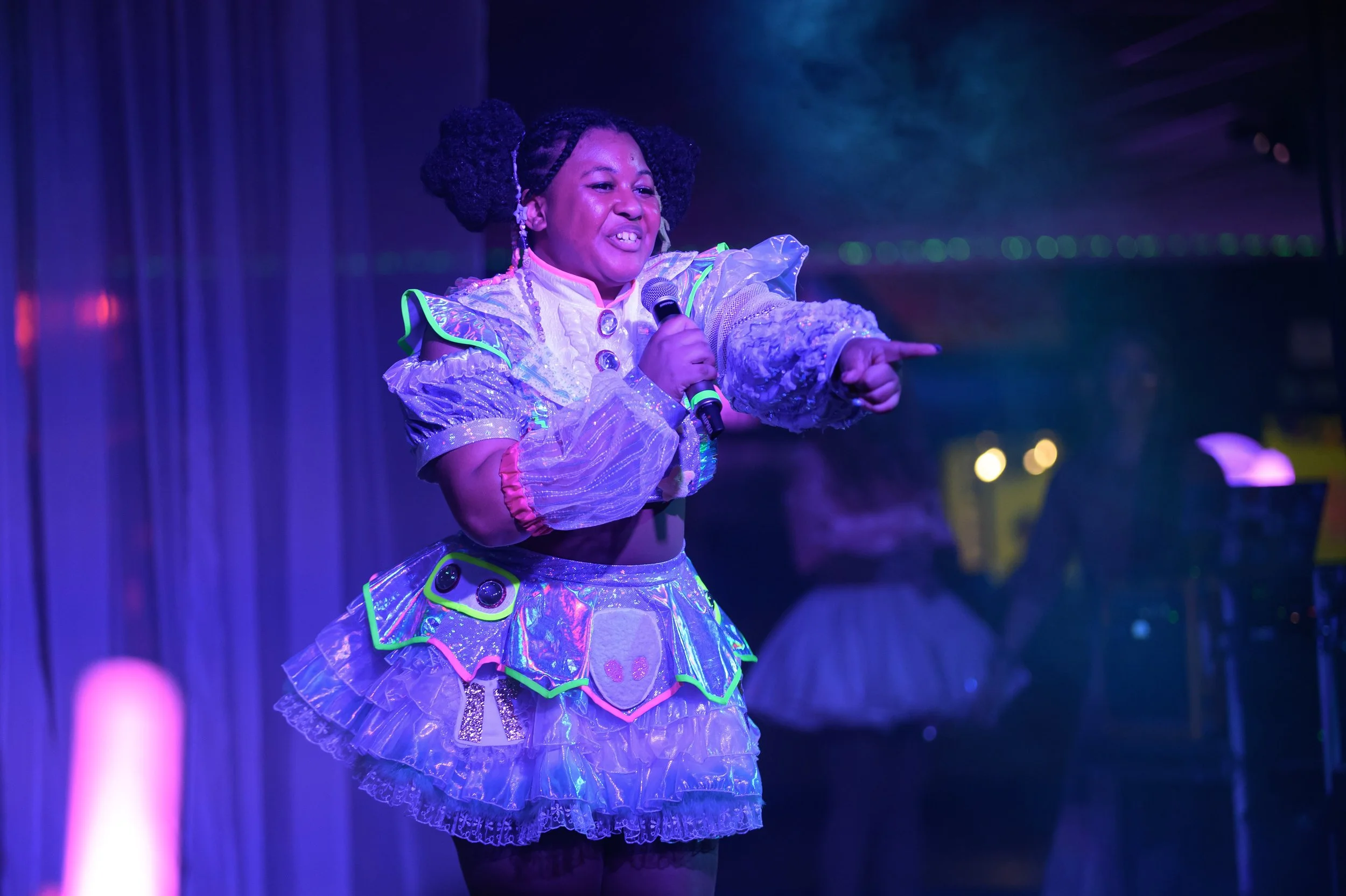 A woman in a colorful, shiny, futuristic costume holding a microphone and pointing, performing on stage with vibrant lighting and a dark curtain background.