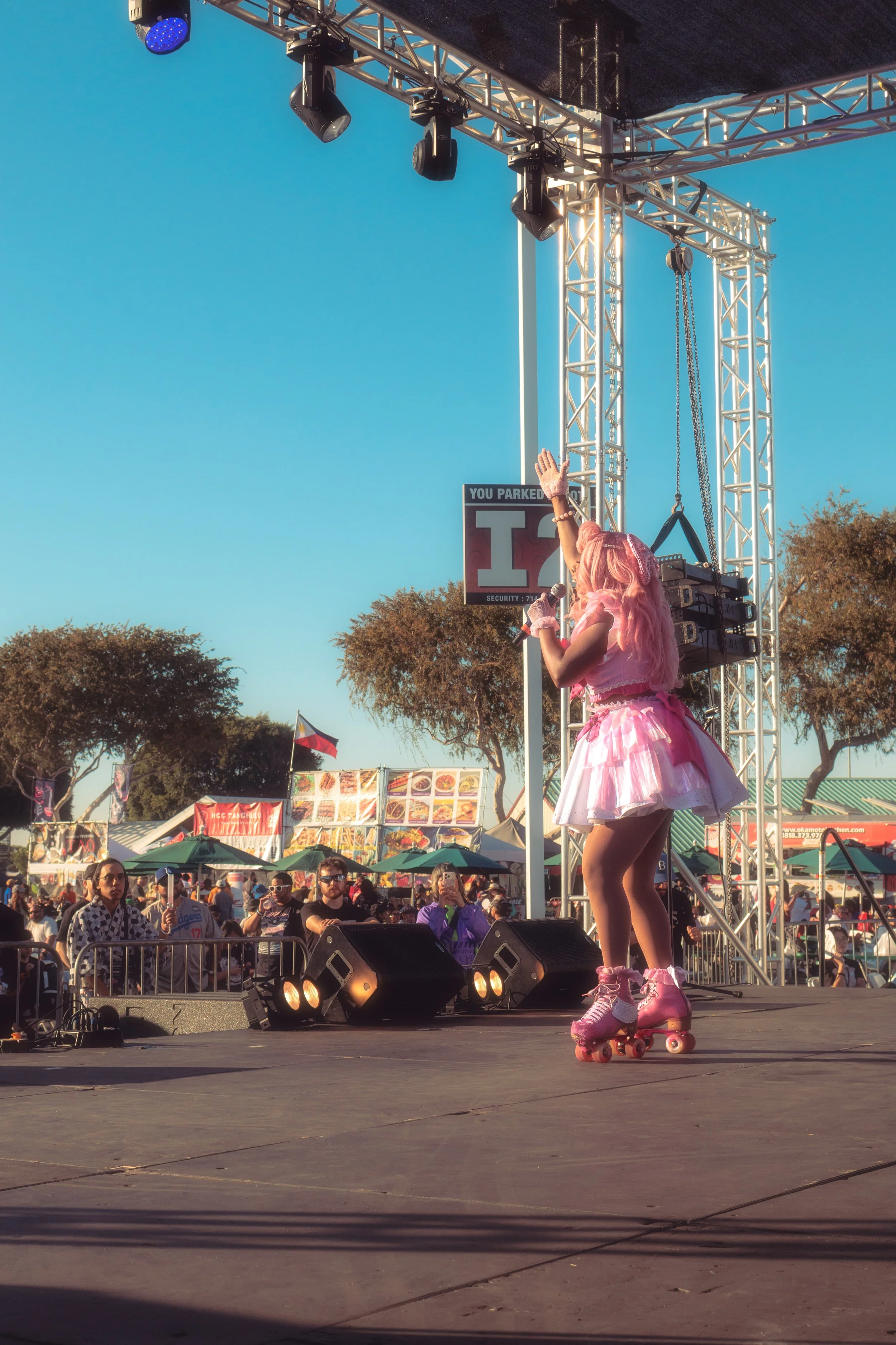 A performer with pink hair, wearing a pink dress and roller skates, on stage at an outdoor festival, singing into a microphone with audience members and food stalls in the background.