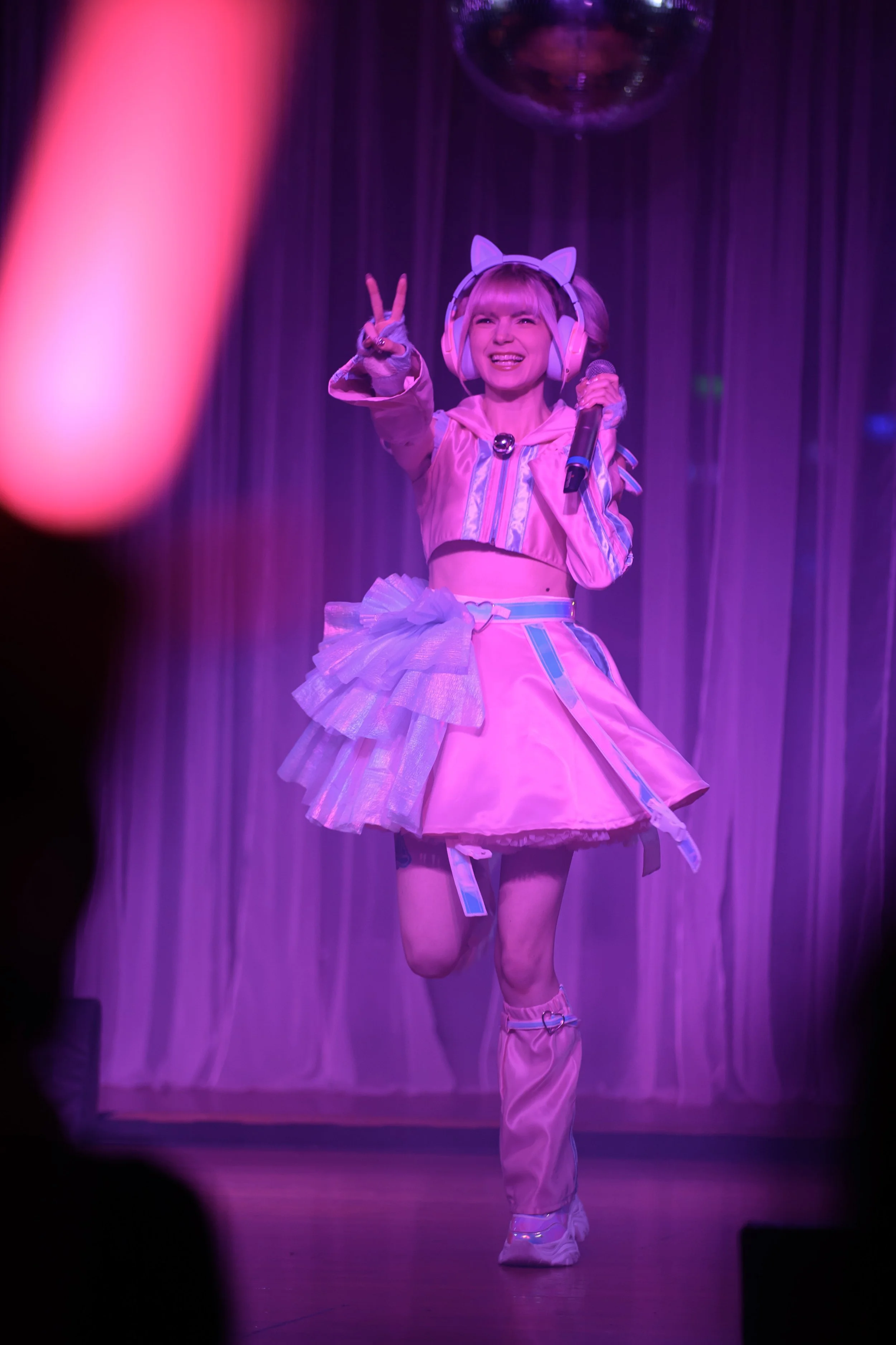A woman in a pink and purple costume with cat ear headphones, holding a microphone, performing on stage with purple lighting and curtains in the background.