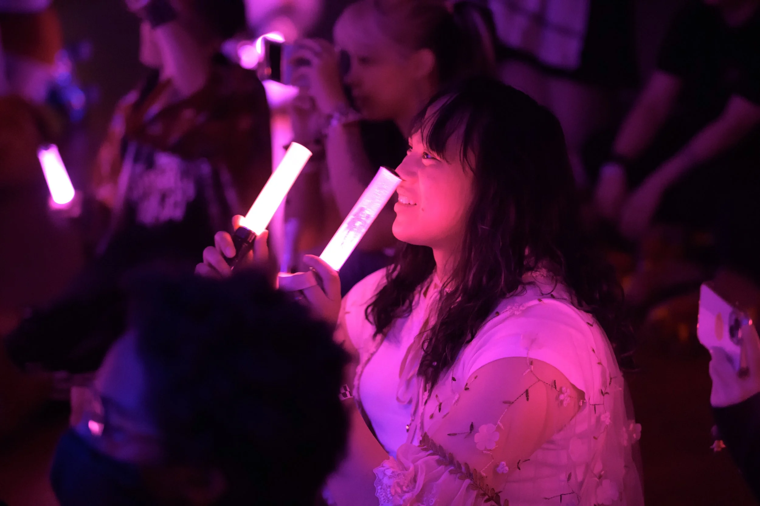 A woman holding a pink glow stick at a concert or event, smiling and enjoying the moment, surrounded by other people with some also holding glow sticks, with pink and purple lighting.