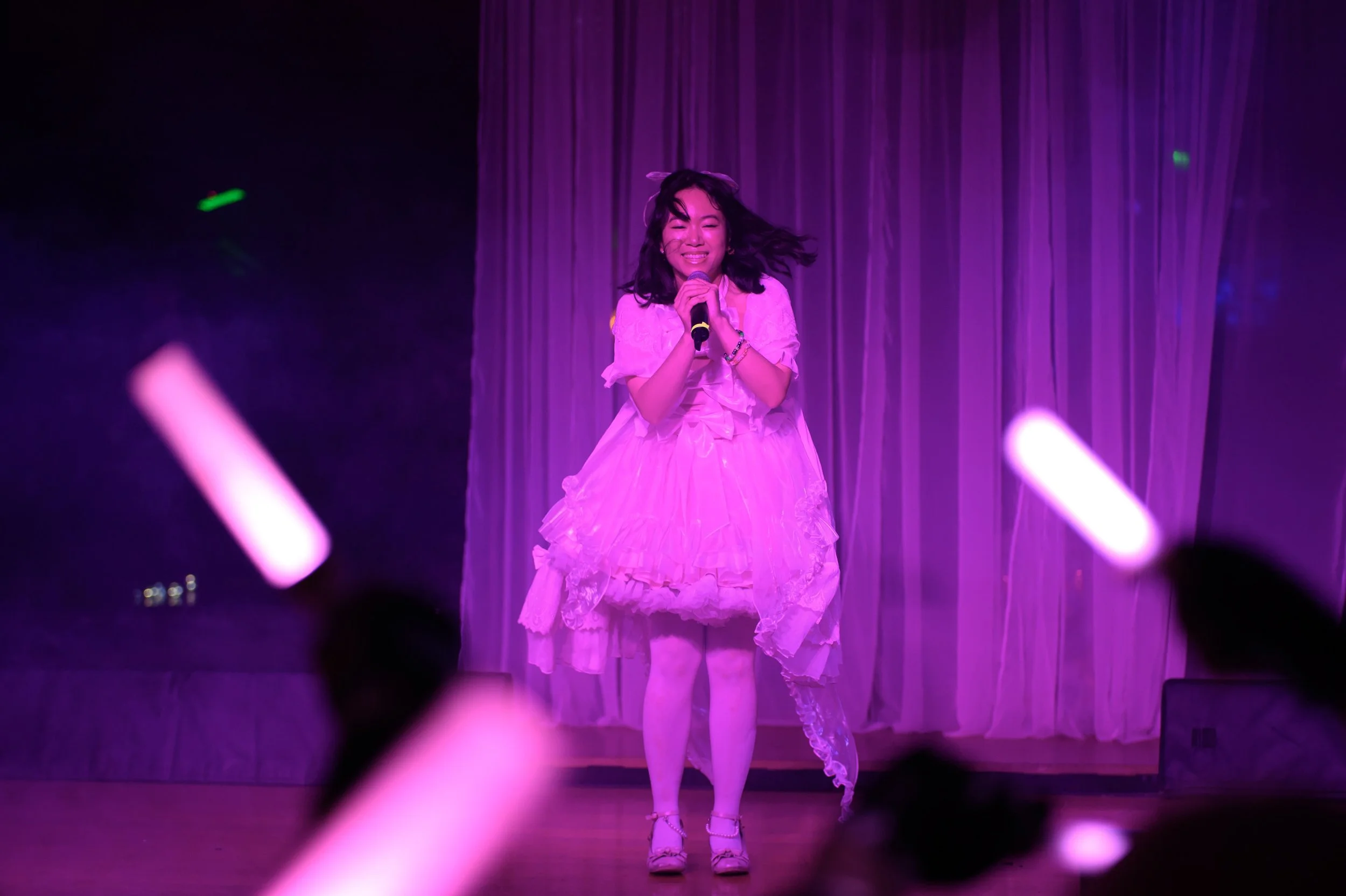 A young woman on stage wearing a pink frilly dress, singing into a microphone, with pink and purple lighting and curtains behind her, audience members holding pink glow sticks.