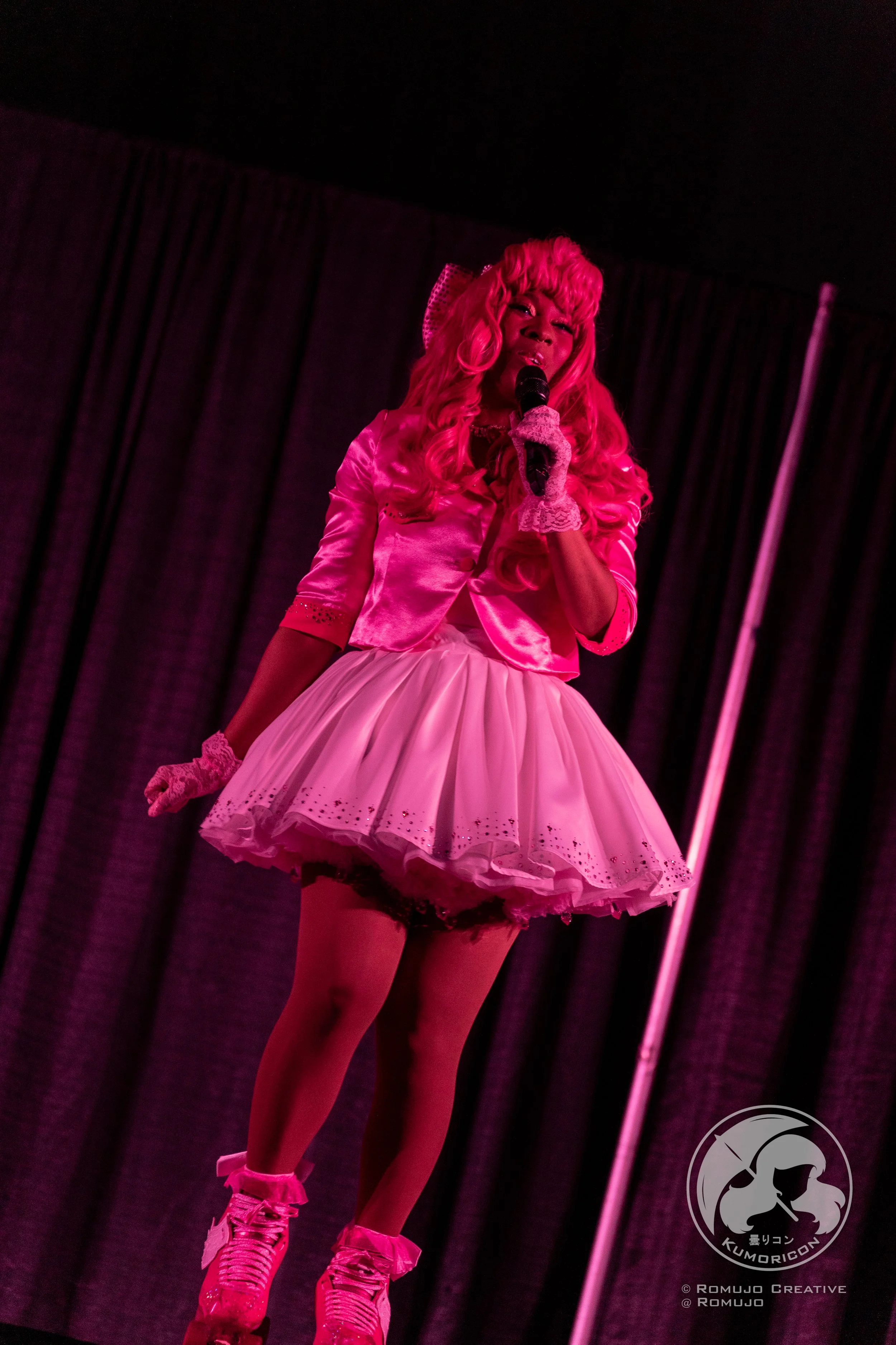 Performer dressed in pink costume singing into microphone on stage with dark curtains background.