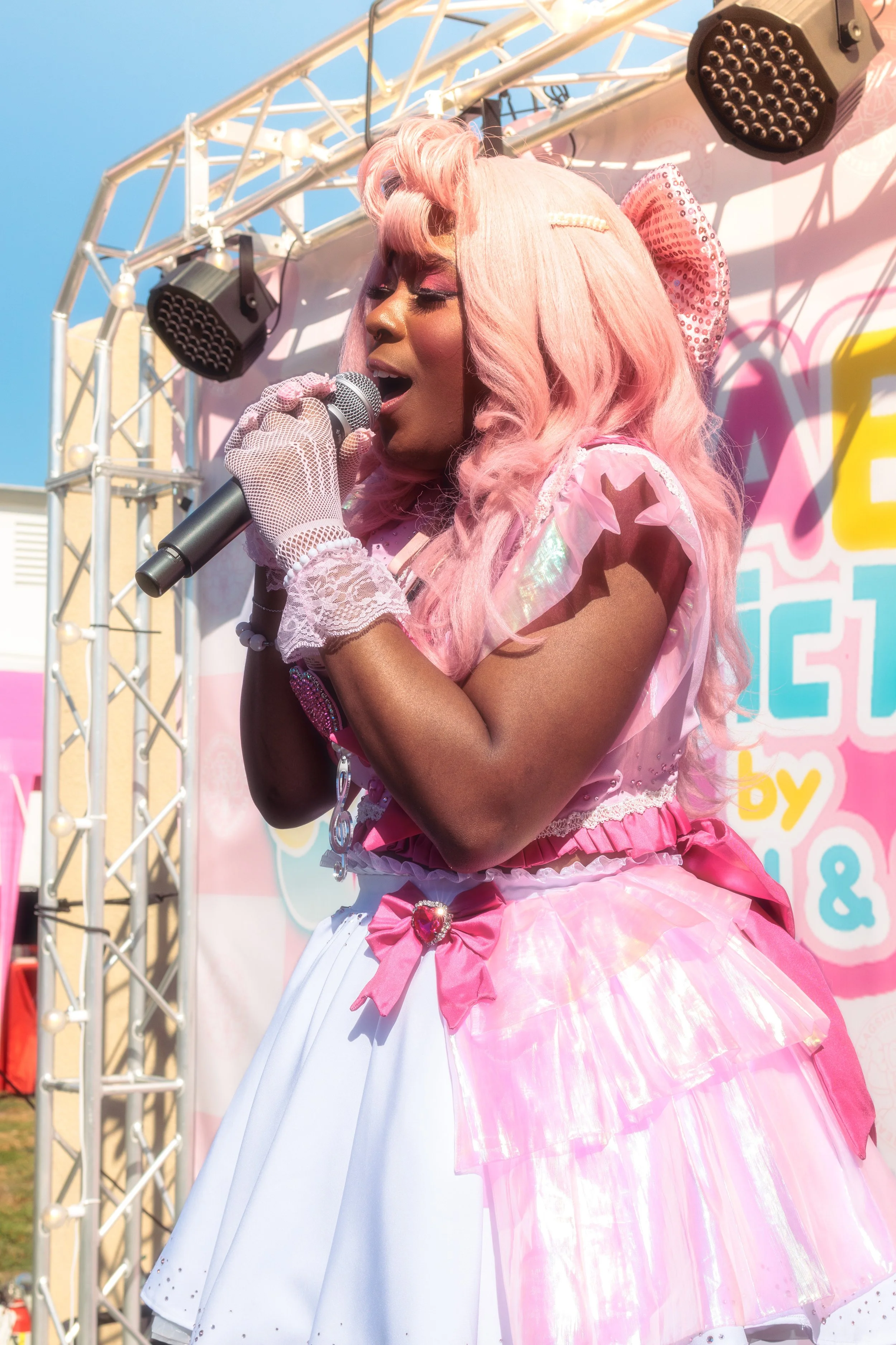 A person with light pink hair styled in curls, singing into a microphone while wearing a pink and white outfit with lace and bows, standing on an outdoor stage with a colorful backdrop.