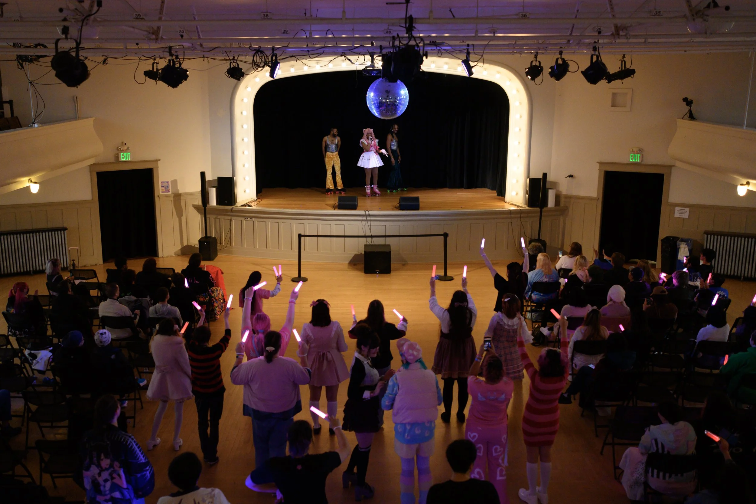 People watching a stage performance in a theater; some audience members are dancing with glow sticks, and performers are dressed in colorful costumes on stage with a disco ball overhead.