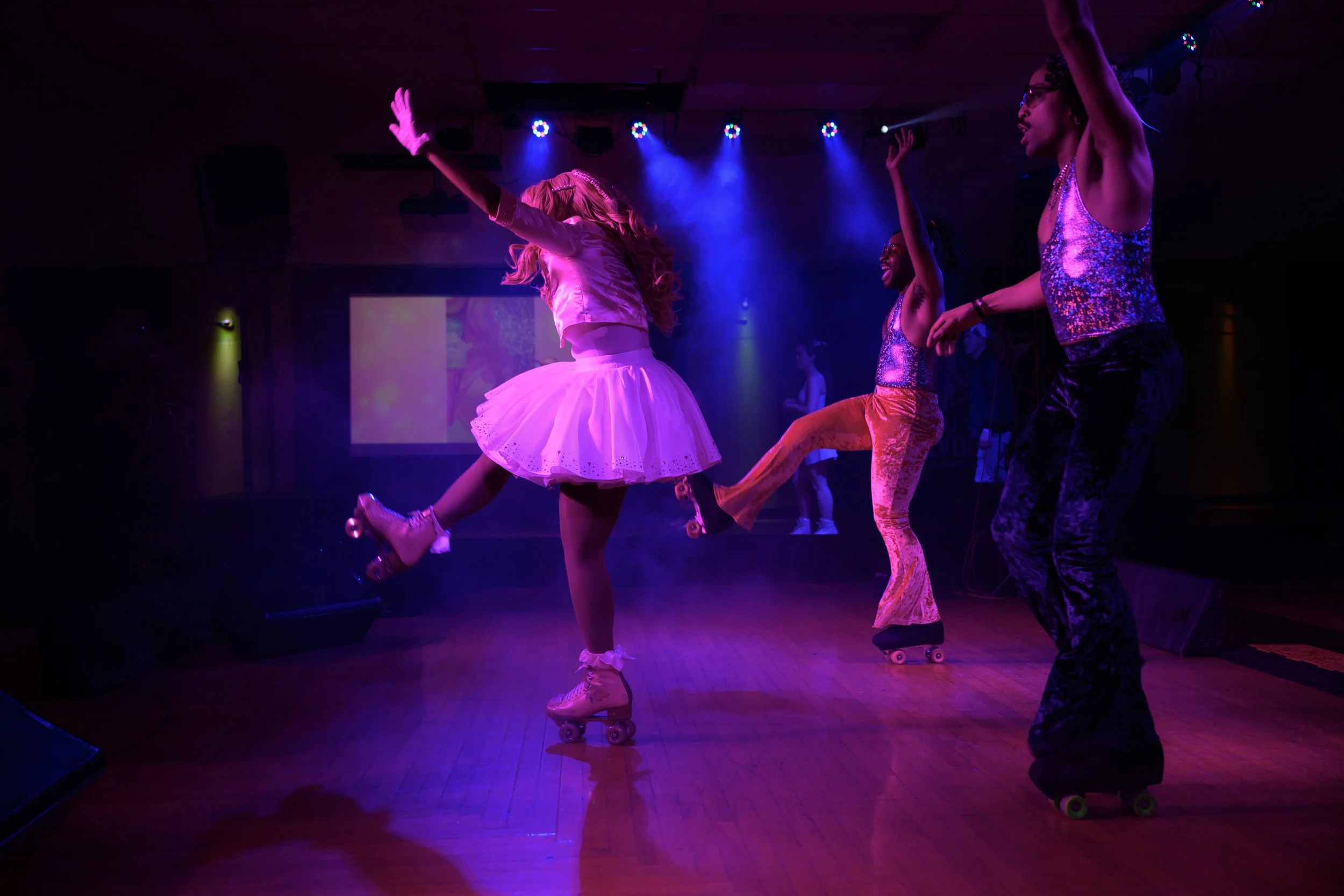 Four women roller skating on a stage with colorful lights, dressed in bright, shiny costumes and posing in dance positions.