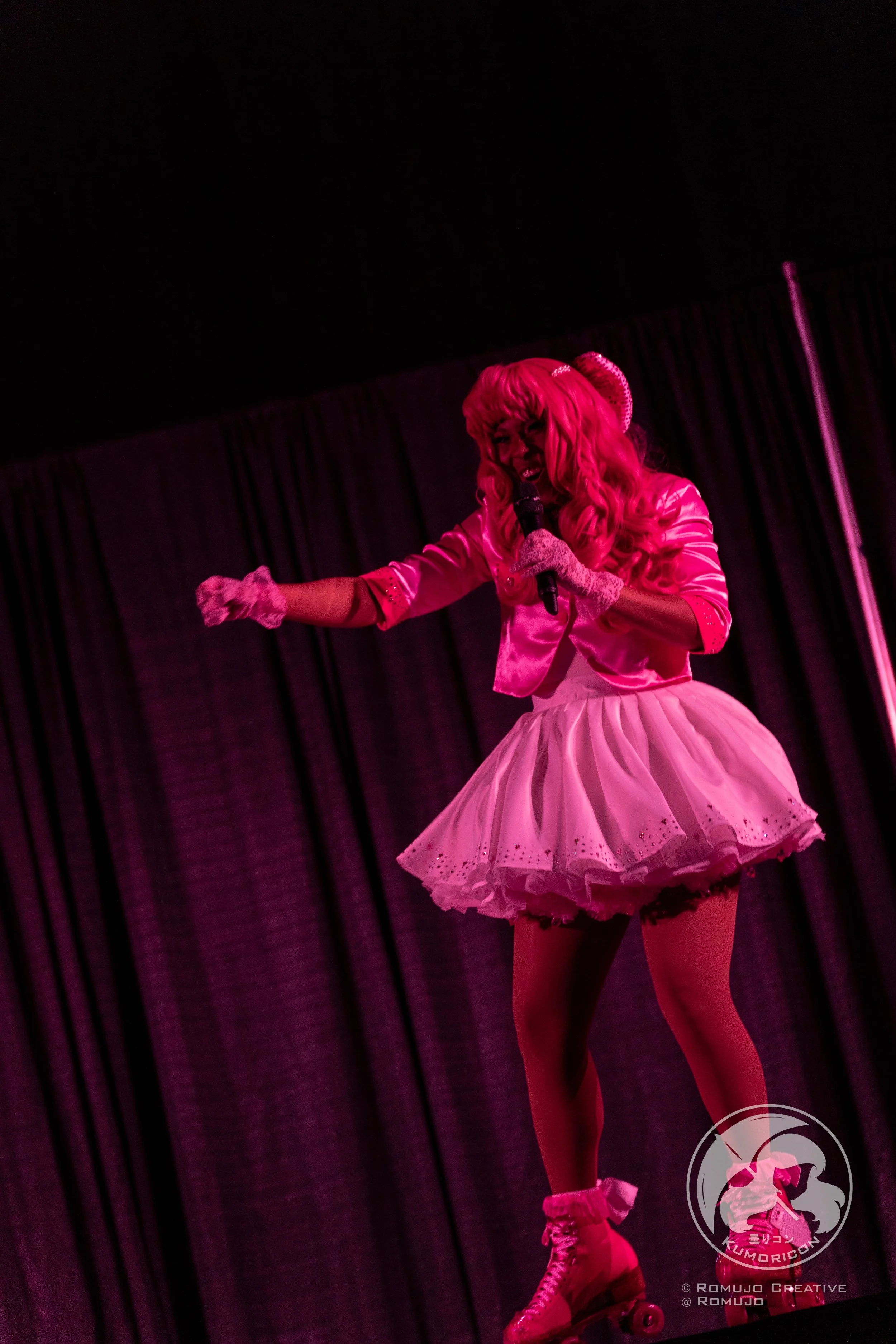 Person dressed in a pink outfit performing on stage with a microphone, in front of black curtains.