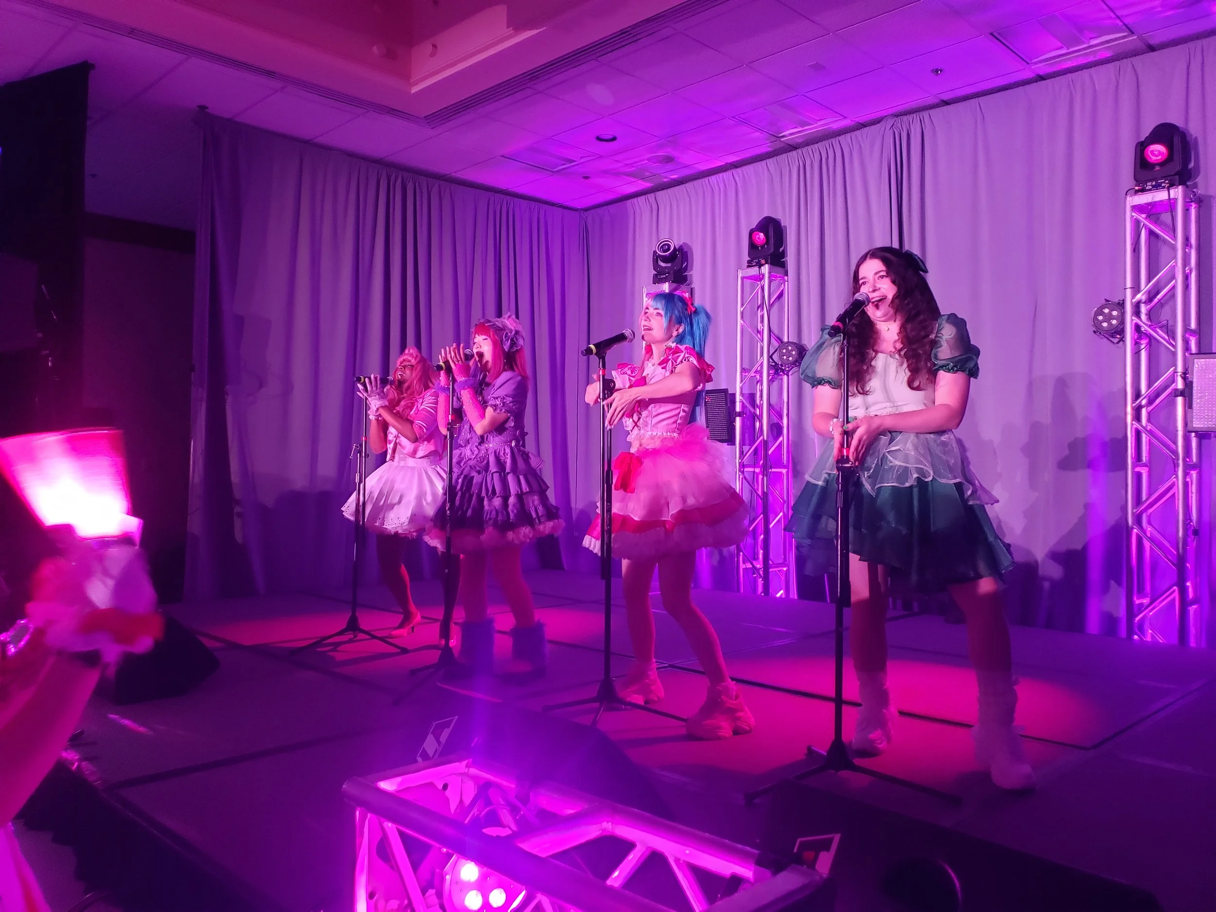 Four performers on stage dressed in colorful, whimsical costumes with tutus and accessories, singing into microphones at a concert or event, illuminated by purple stage lights with a backdrop of curtains and stage lighting equipment.