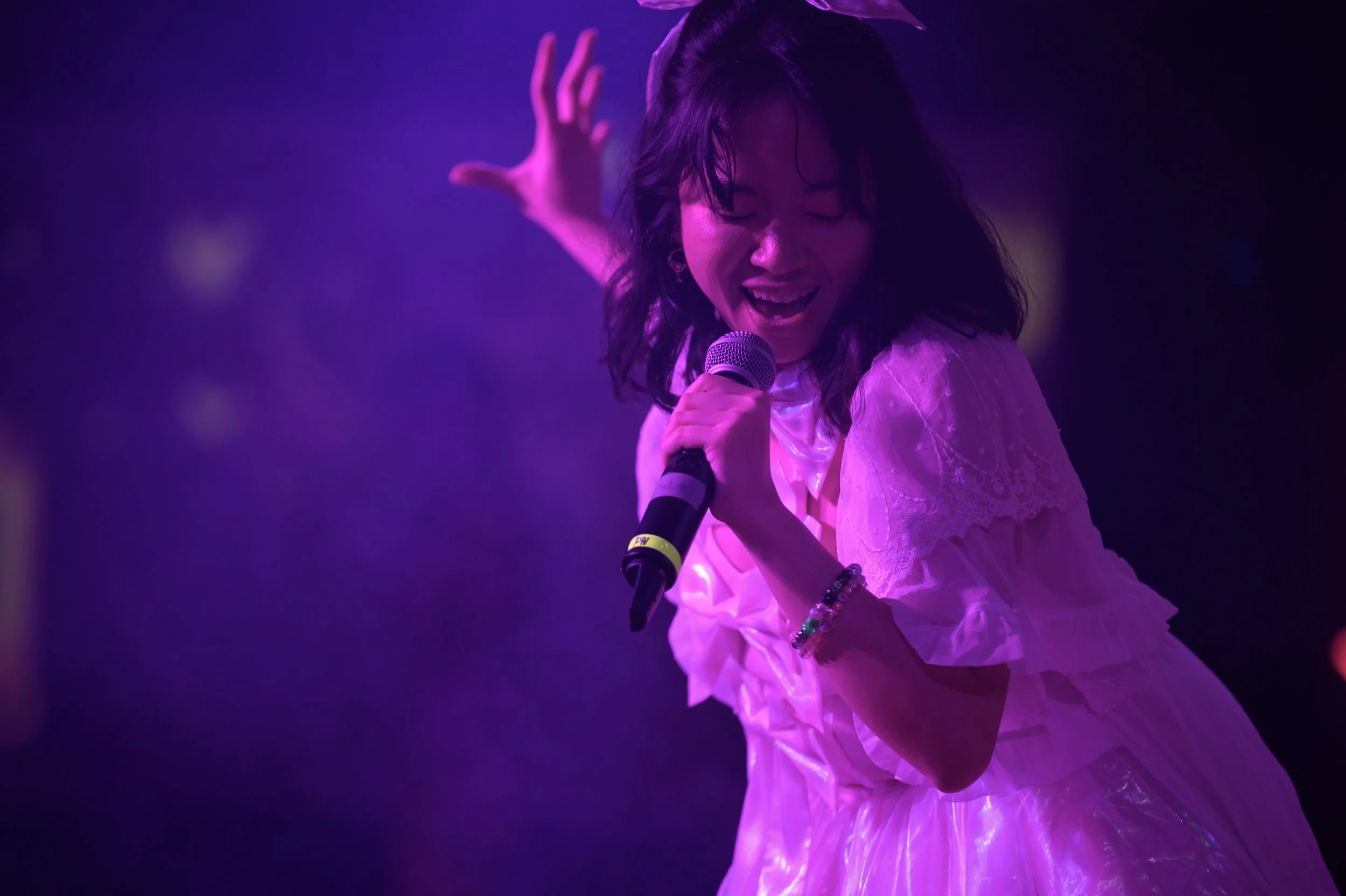 A woman singing into a microphone on stage, illuminated by purple lighting. She is wearing a white dress with lace details and has shoulder-length dark hair.