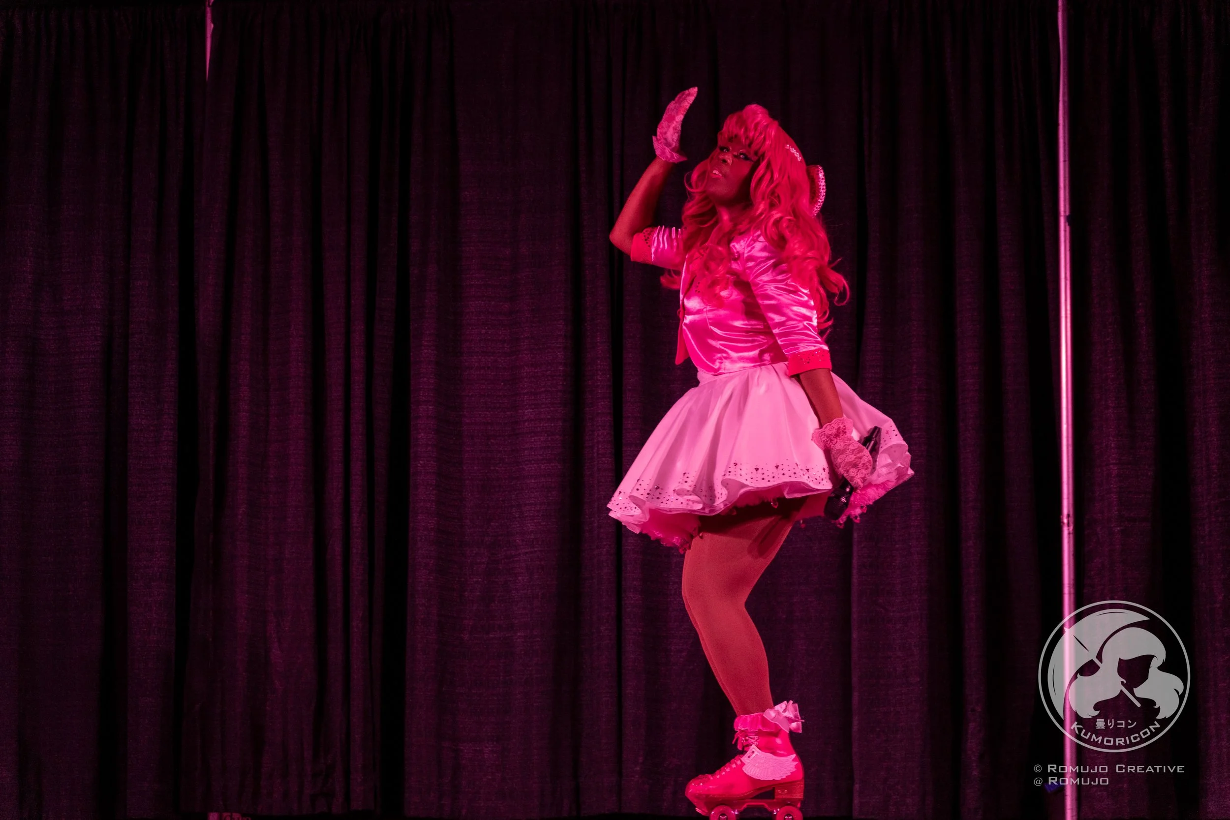 A person dressed in a pink costume with a tutu skirt, roller skates, and gloves, striking a pose on stage with a dark curtain background.