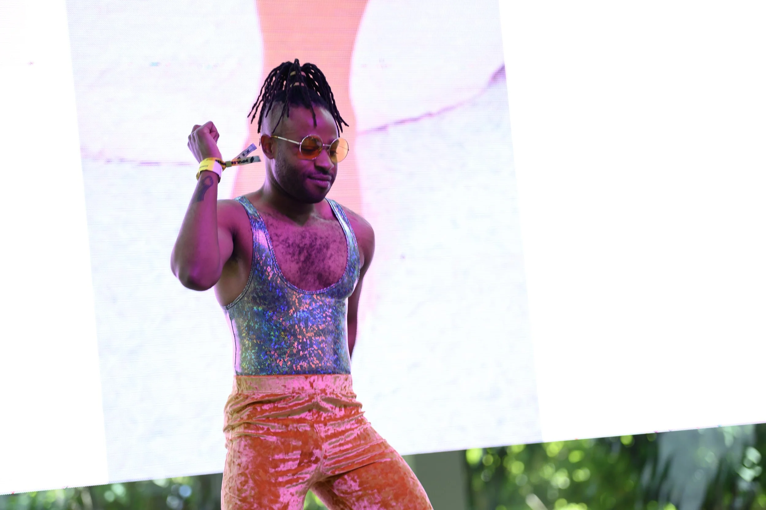 Person with dreadlocks wearing iridescent tank top, gold velvet pants, and sunglasses, dancing against a bright screen background.