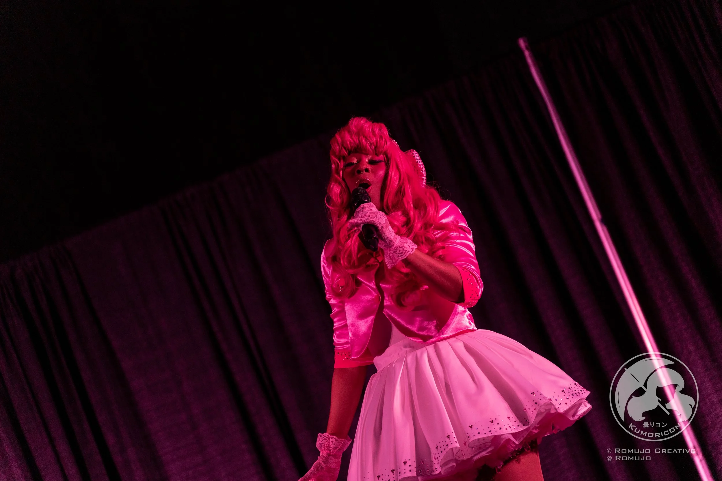 Performer with pink hair and pink satin outfit singing on stage, with black curtain background and pink lighting.