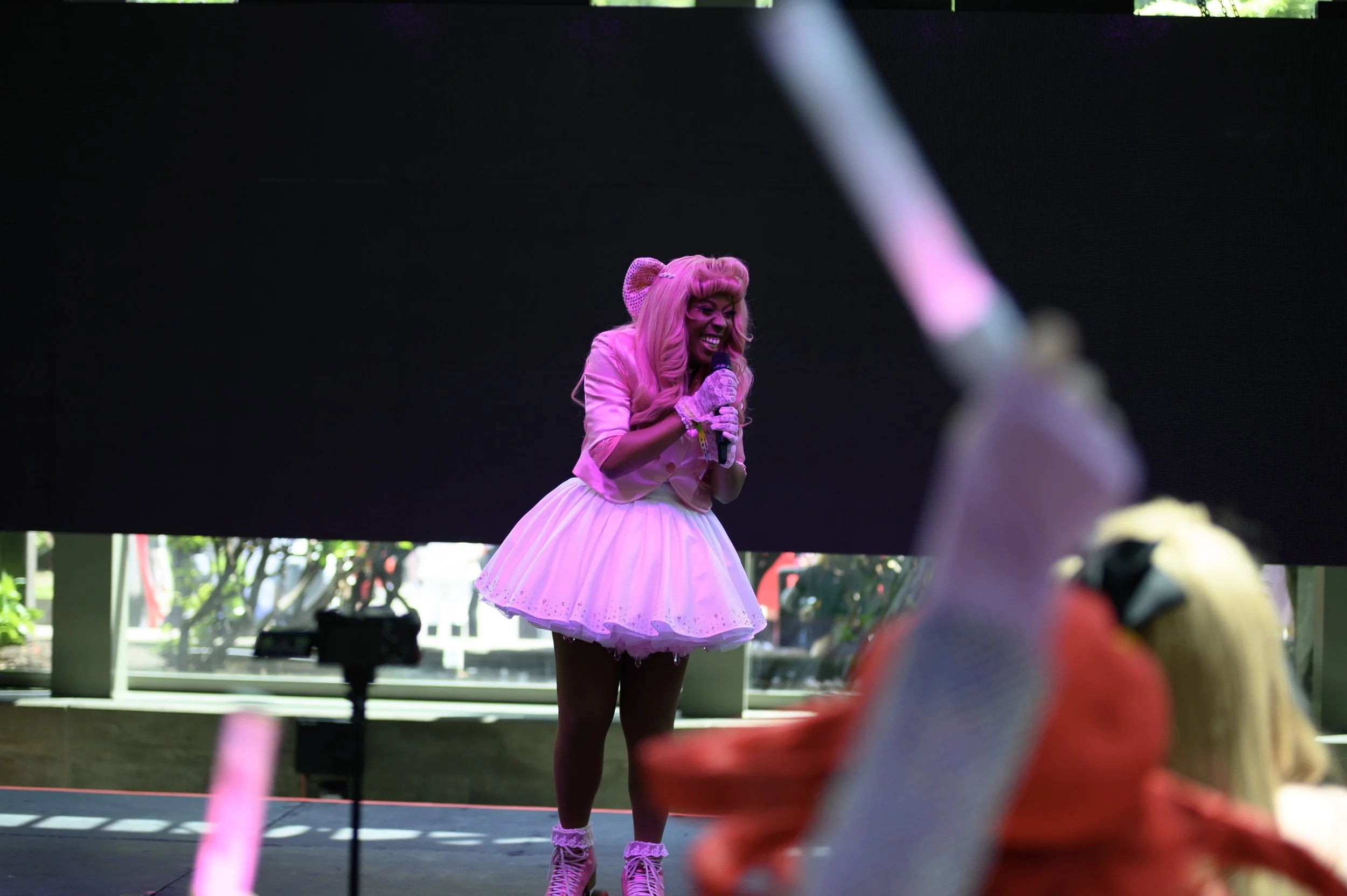 A woman dressed in a pink wig, pink gloves, and a pink tutu skirt performing on stage, holding a microphone with a big smile.