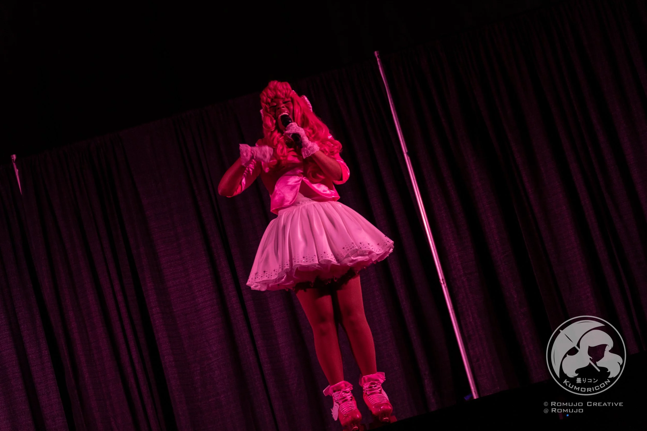 Performer on stage in pink costume, singing into a microphone, with a dark curtain background.