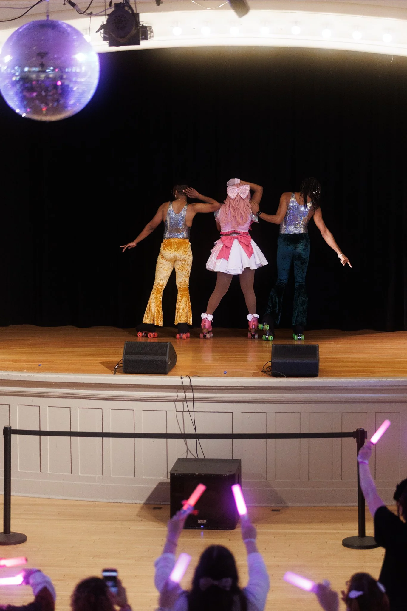 Three girls are roller skating on stage, dressed in colorful costumes, performing in front of an audience with glow sticks.