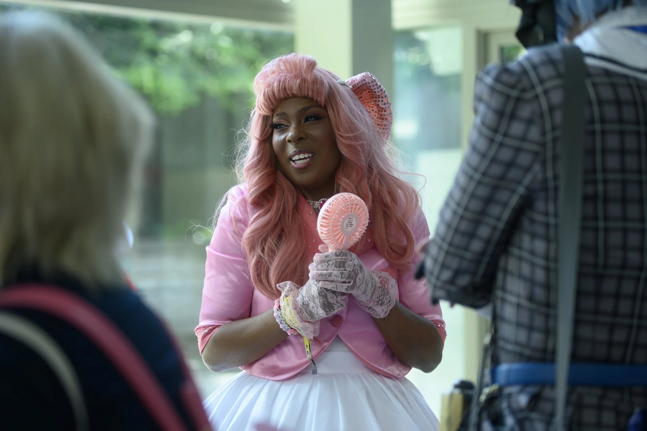 A woman with pink hair, dressed in a pink jacket, white dress, and lace gloves, holding a small pink fan and talking to two other people at an indoor location with large windows and greenery outside.