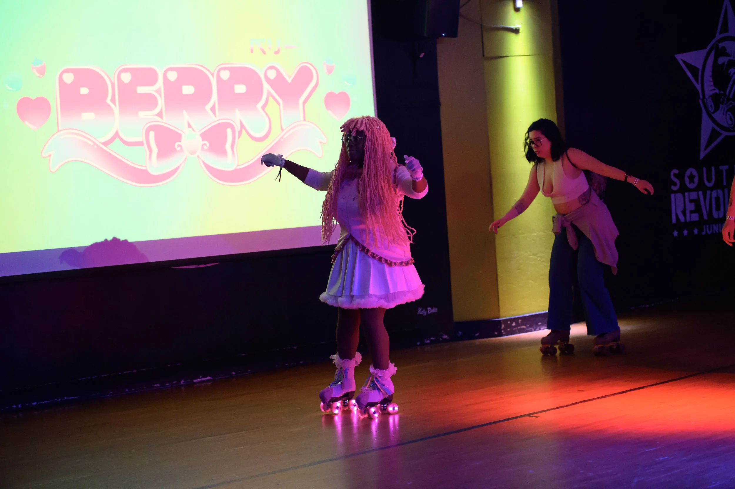 Two women roller skating at a brightly lit indoor event, with a large colorful screen displaying the word 'BERRY' in the background.