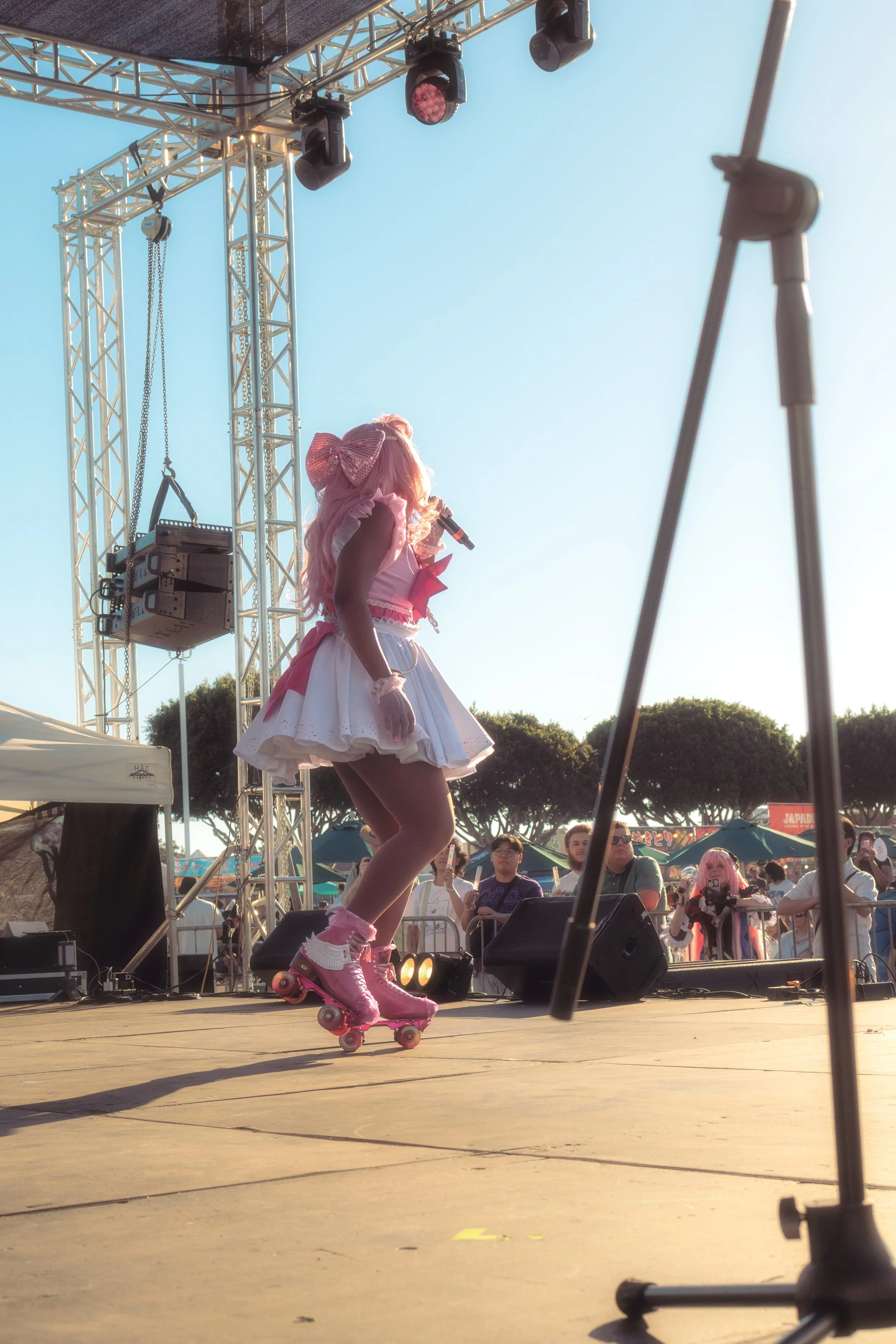 A woman dressed in a pink and white costume with a bow and roller skates performing on an outdoor stage during daytime. The audience and trees are visible in the background.