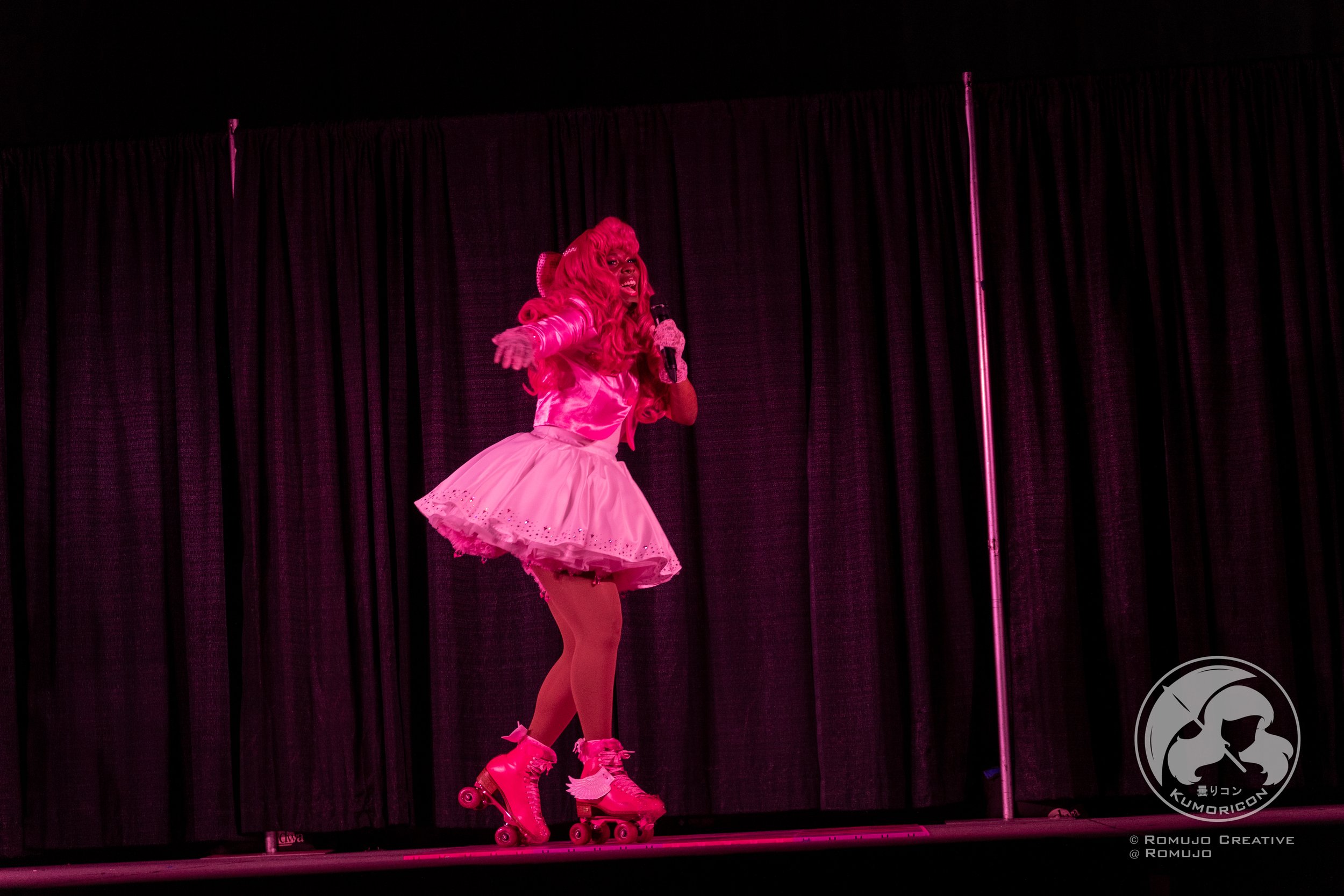 Performer dressed in pink with roller skates singing on stage in front of black curtains.