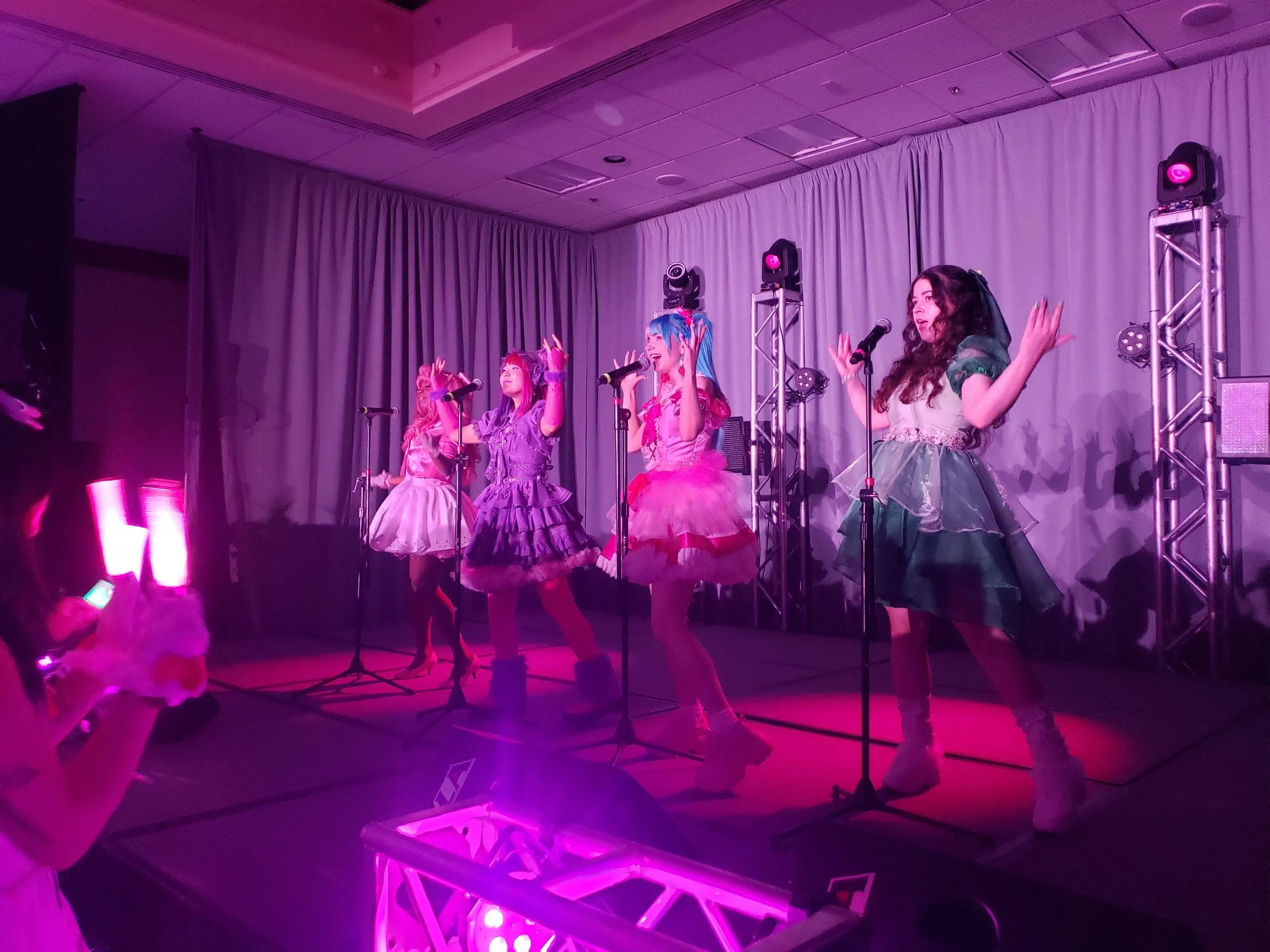 Four female performers dressed in colorful, idol costumes performing on stage with purple lighting and gray curtains in a room with a low ceiling.