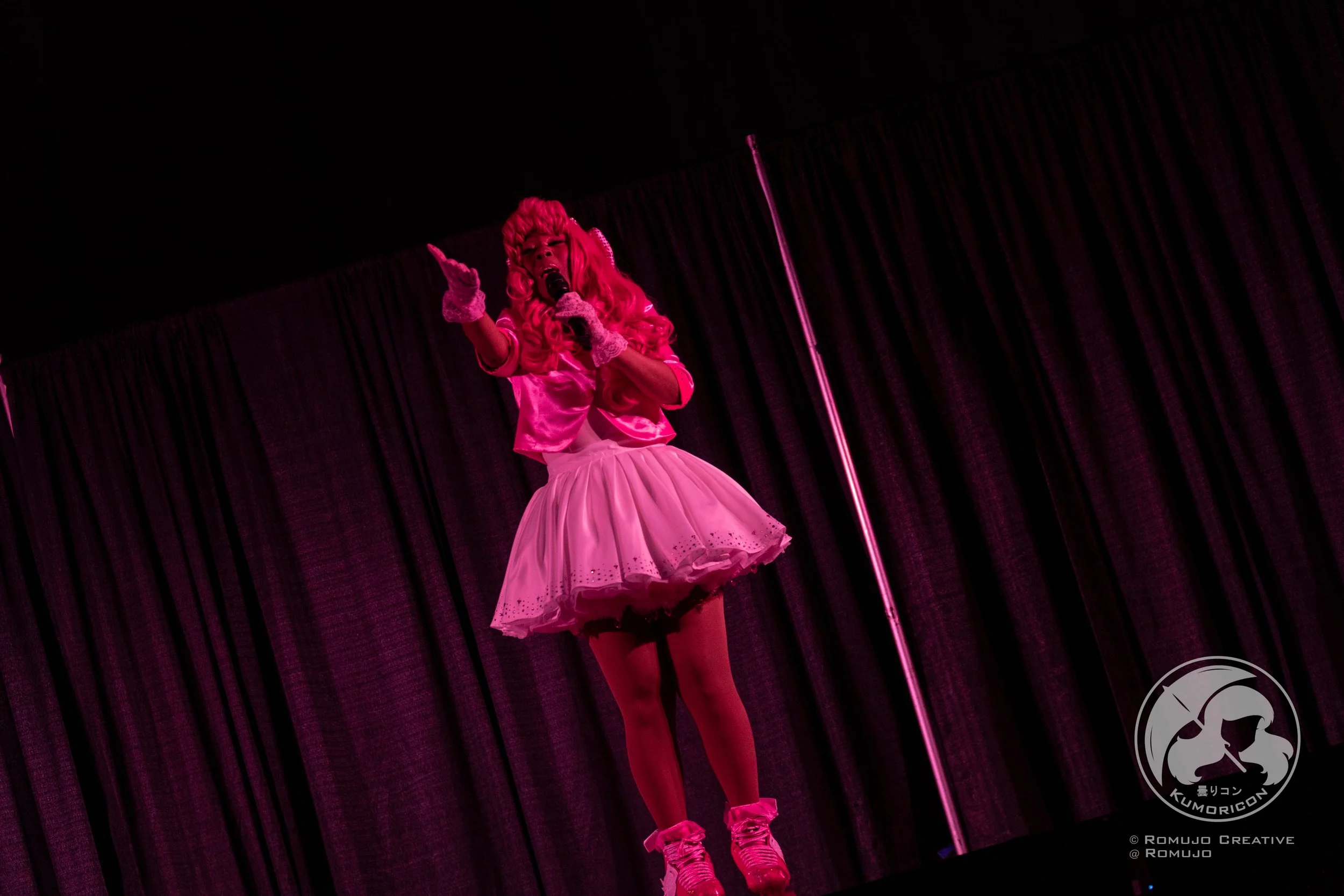 Performer dressed in a pink costume with a fluffy skirt, pink stockings, and pink shoes on stage, singing into a microphone with black curtains in the background.