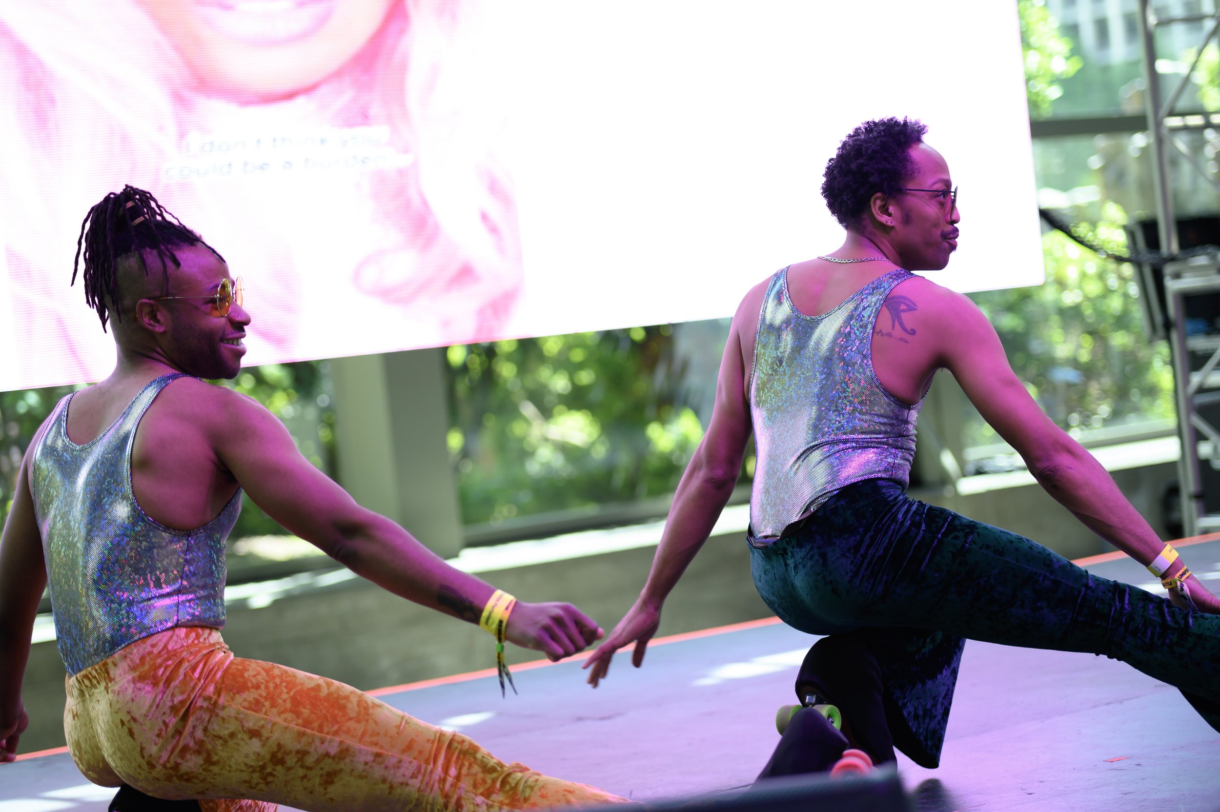 Two people performing a skate move on stage, hands down while leaning back on one knee, with a large screen behind them and greenery outside visible through windows.