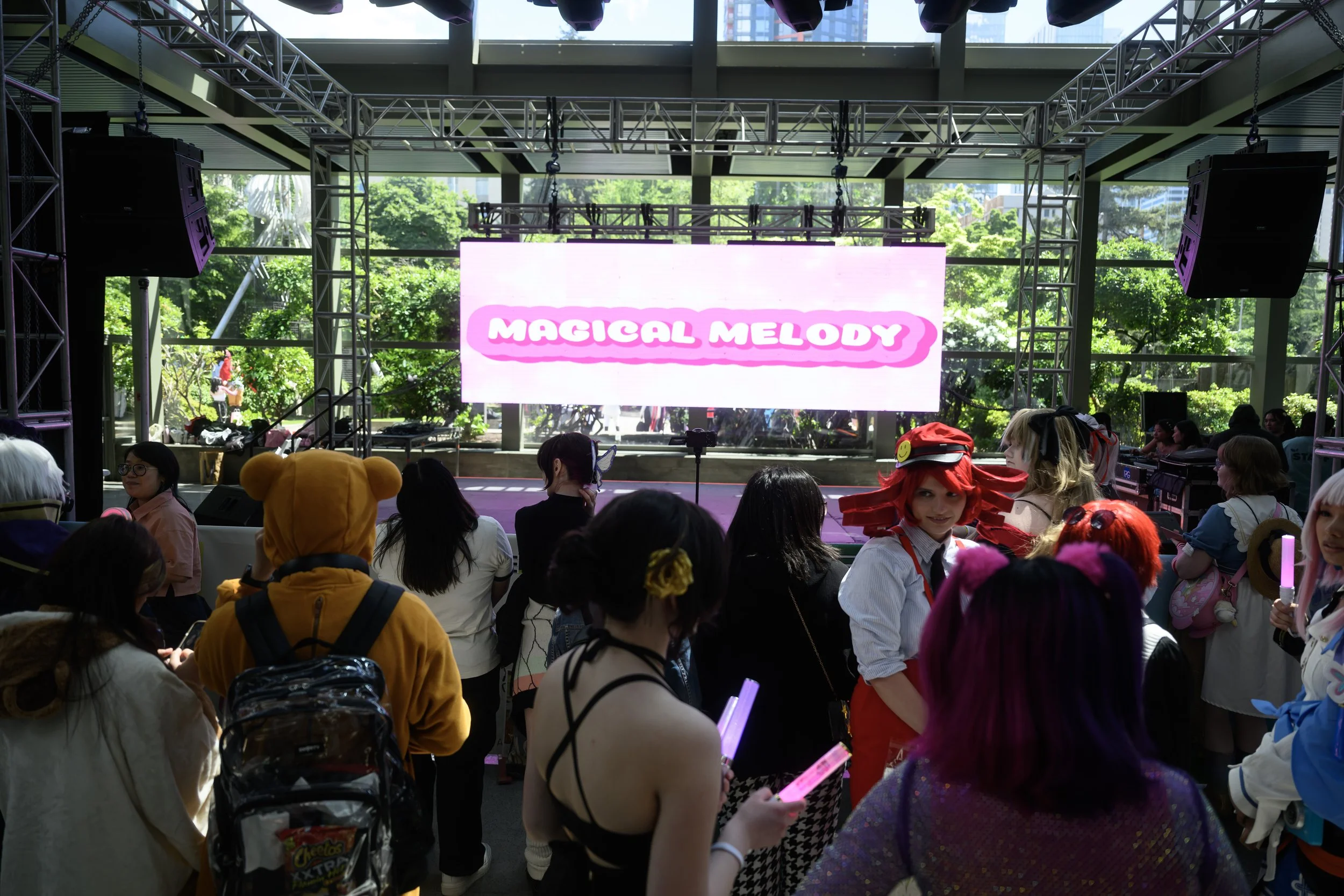 Indoor stage setup with a large digital screen displaying "MAGICAL MELODY," surrounded by a crowd of people, some in colorful costumes, in a brightly lit space with large windows showing greenery outside.