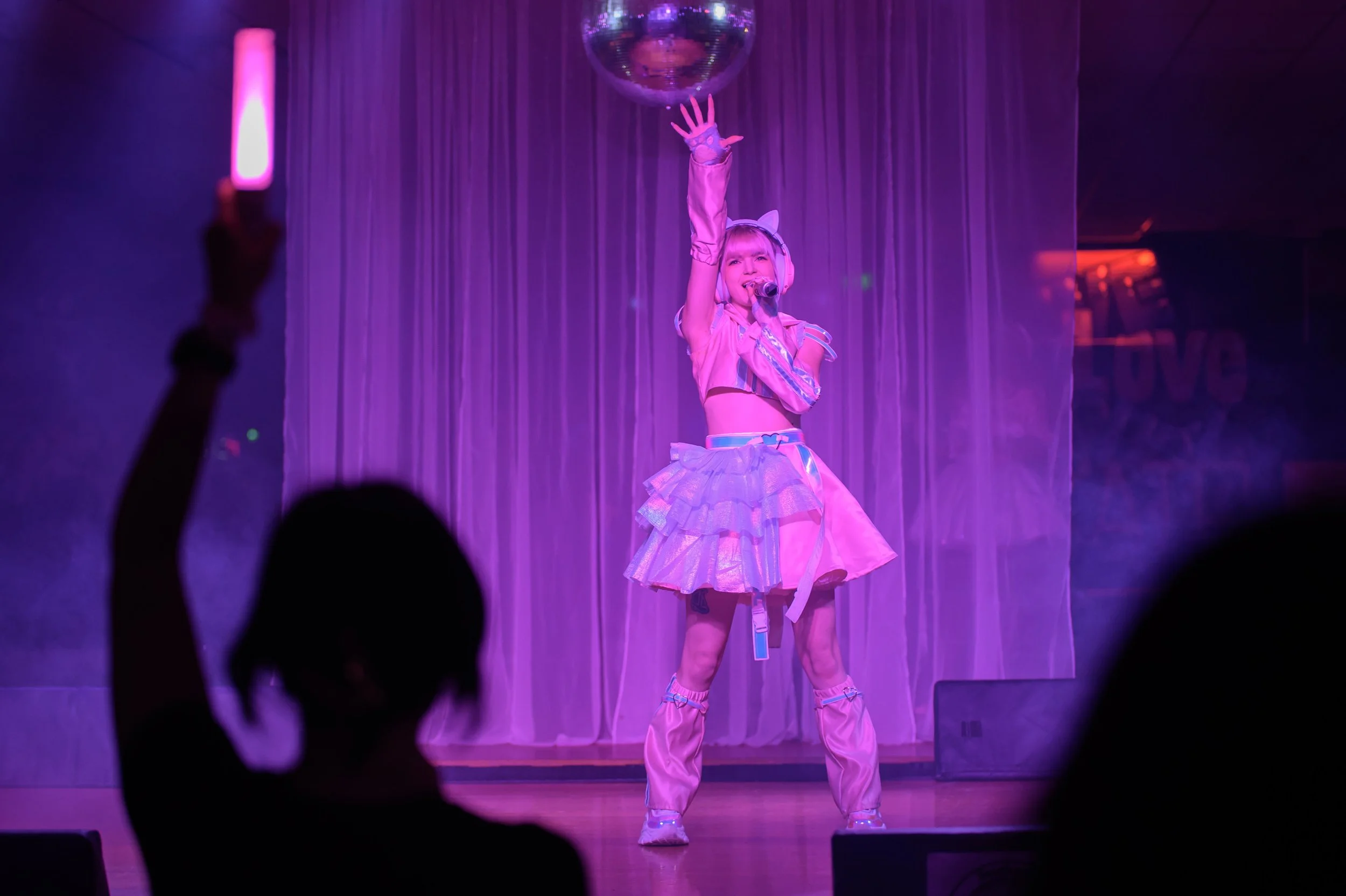 A performer dressed in a pastel-colored, fantasy-like costume with cat ears, singing into a microphone on stage. The stage is lit with purple and pink lighting, and a disco ball hangs above. Audience silhouettes are visible in the foreground.
