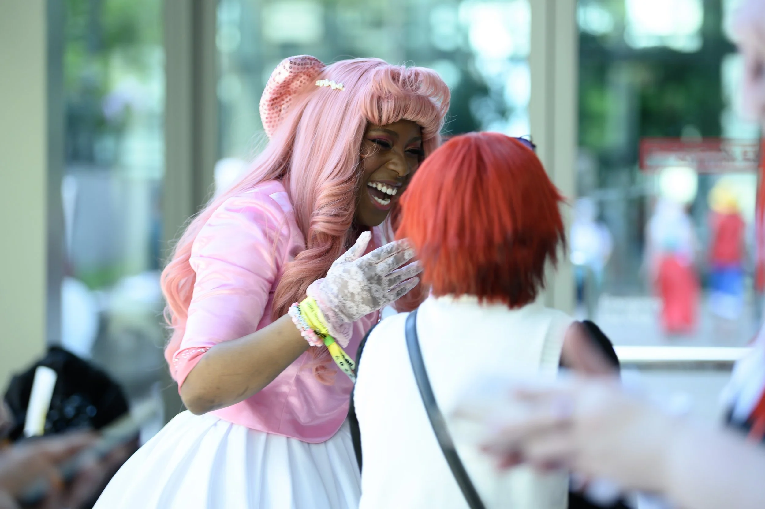 A woman with pink hair, pink dress, and lace gloves smiling and talking to another woman with red hair in a public indoor space.