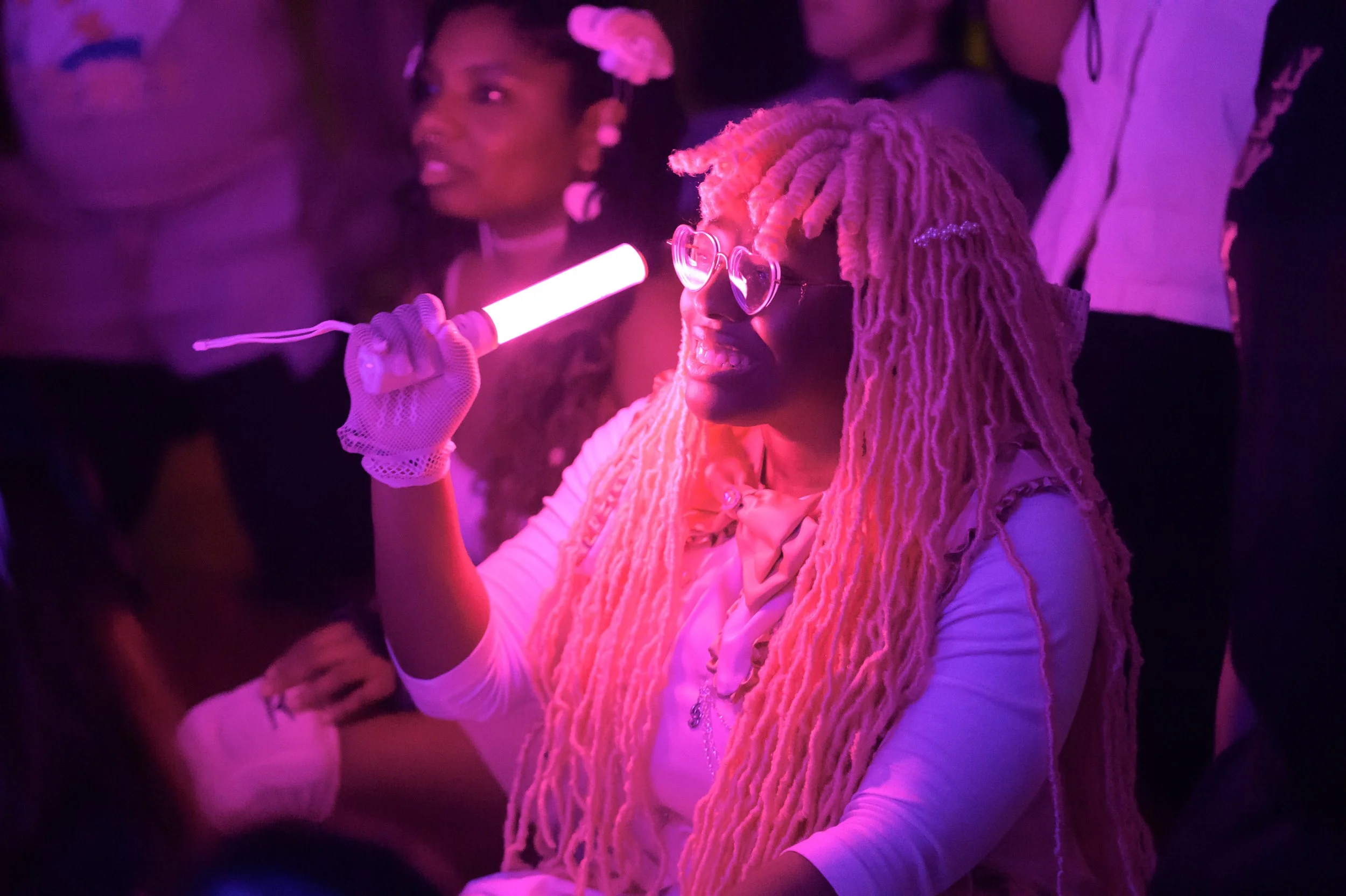 A woman with long pink dreadlocks, glasses, and a white outfit holding a pink glow stick and smiling at a party or concert with purple lighting.