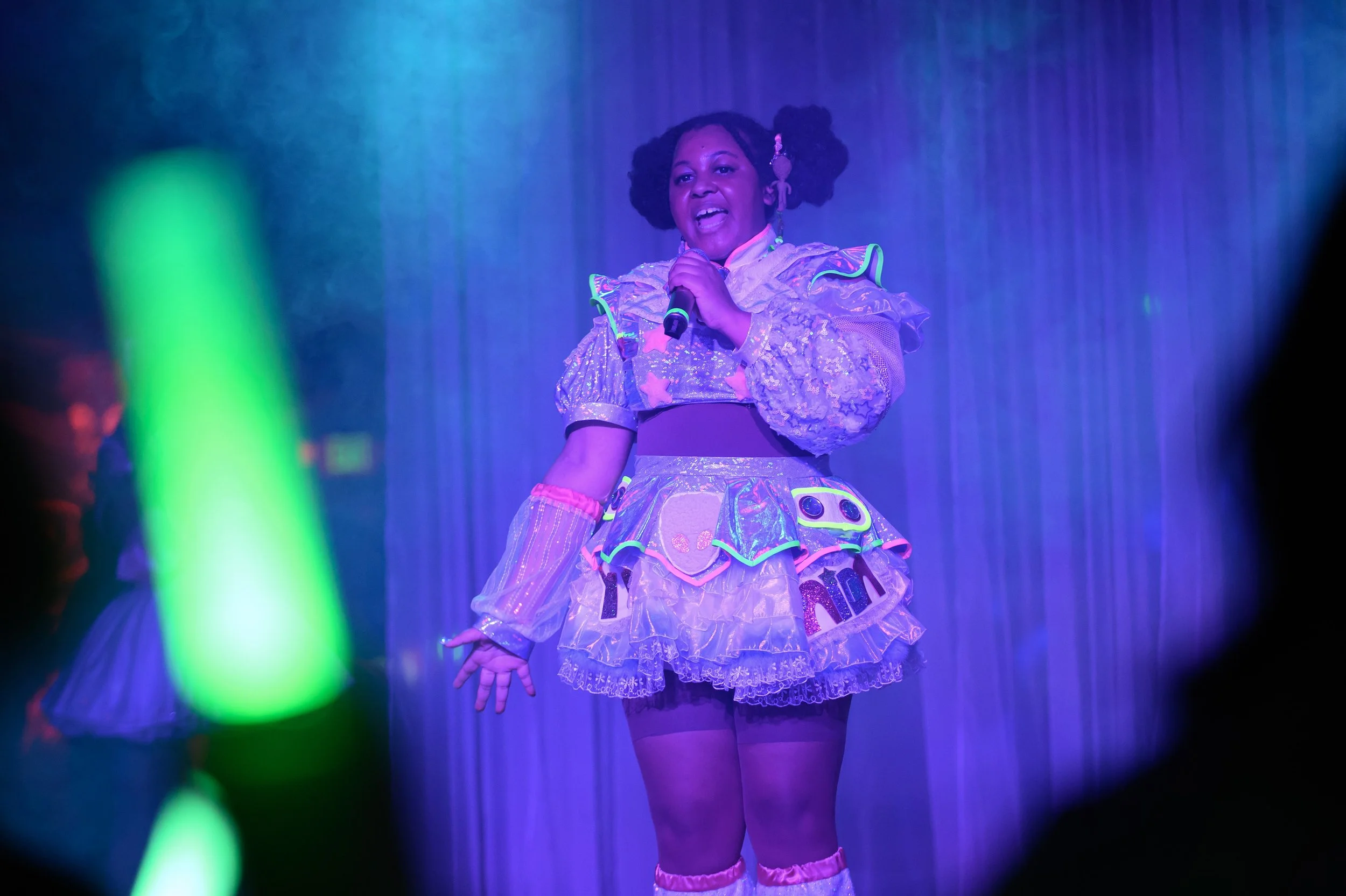 A woman dressed in colorful, shiny, and playful stage costume singing into a microphone on stage with purple curtain background, neon green glow stick partially visible in foreground.