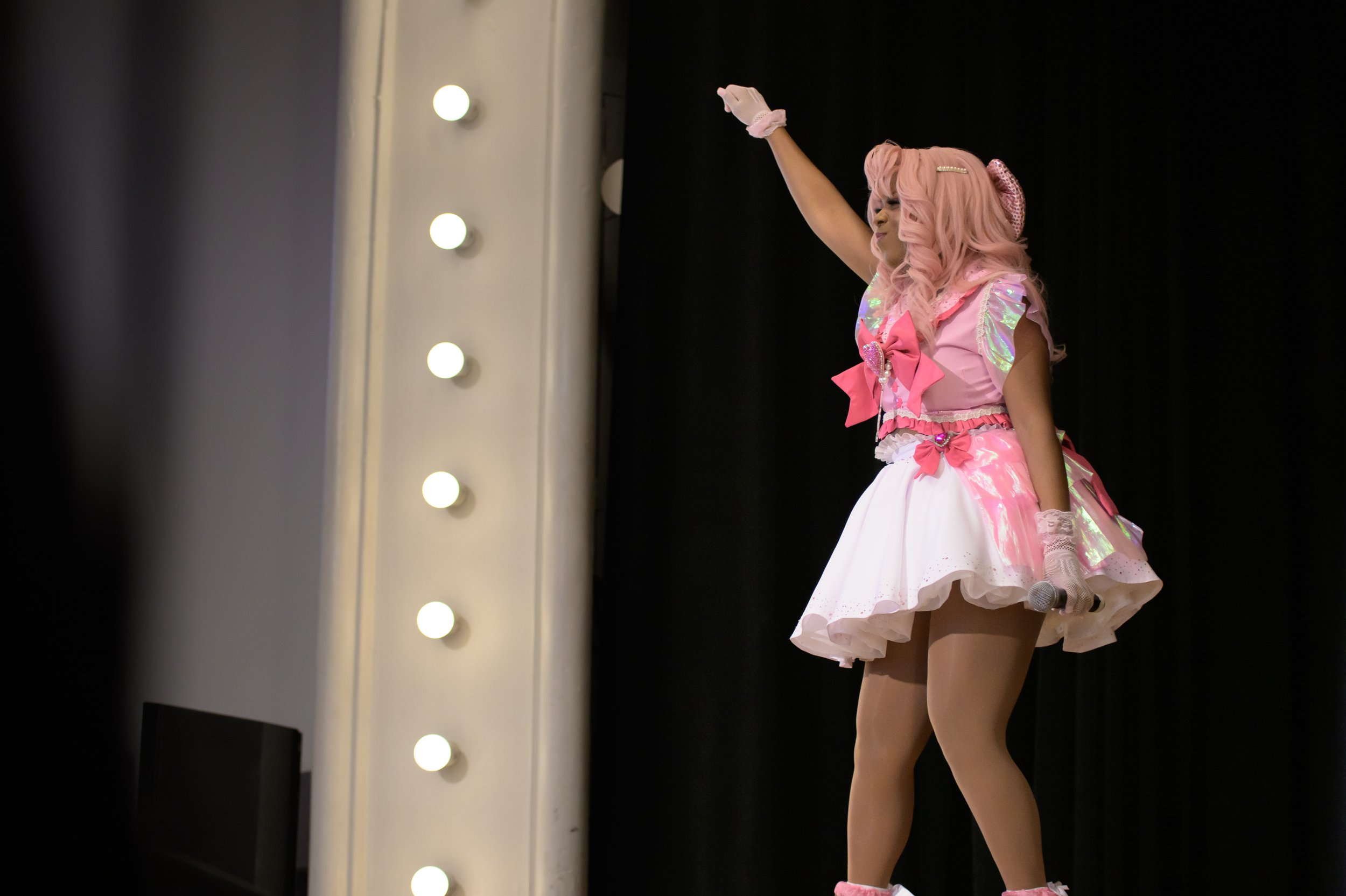 Performer dressed in a pink and white costume with pink hair on stage, waving to the audience, holding a microphone, with black curtains behind.