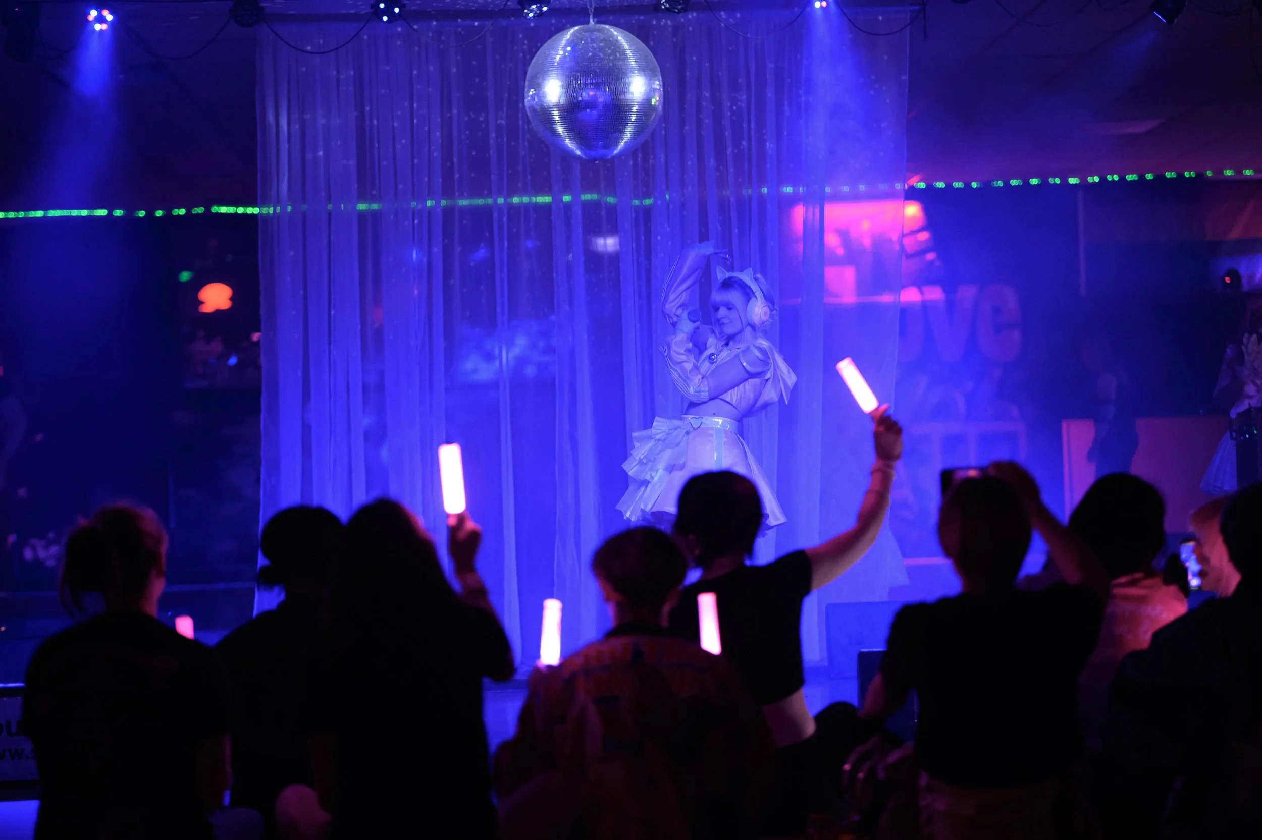 A performer on stage dressed in a futuristic outfit with headphones, standing behind a curtain with purple lighting, and a disco ball overhead. Audience members in the foreground are holding glowing sticks.