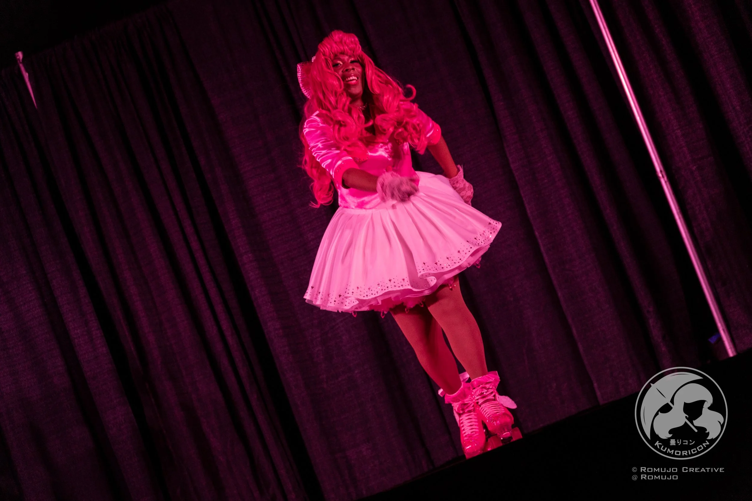 Performer dressed in pink wig, pink dress, and roller skates on stage with black curtain backdrop.