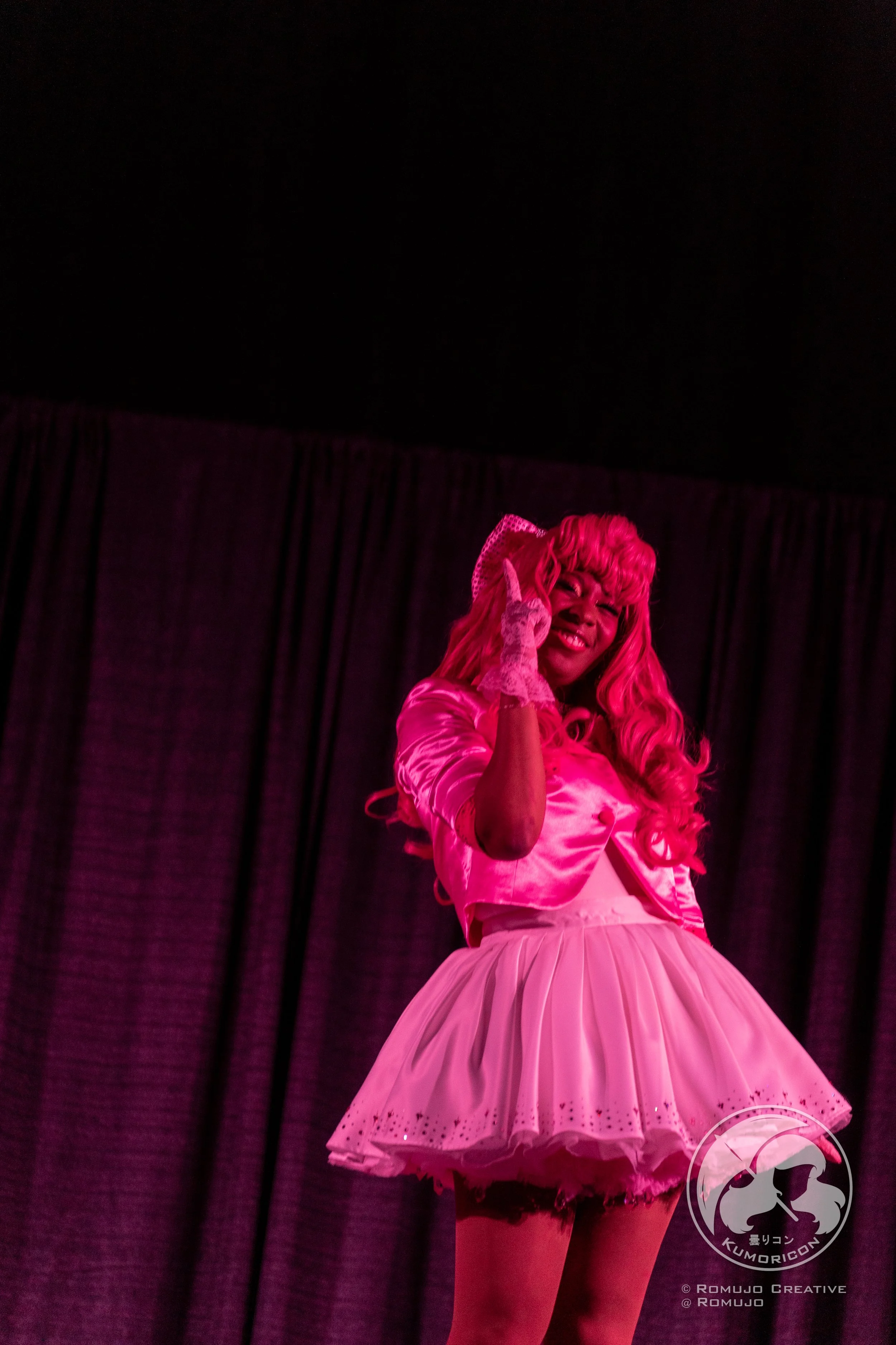 Performer on stage dressed in pink, winking and smiling, with a pink wig and a frilly pink skirt, standing in front of a dark curtain.
