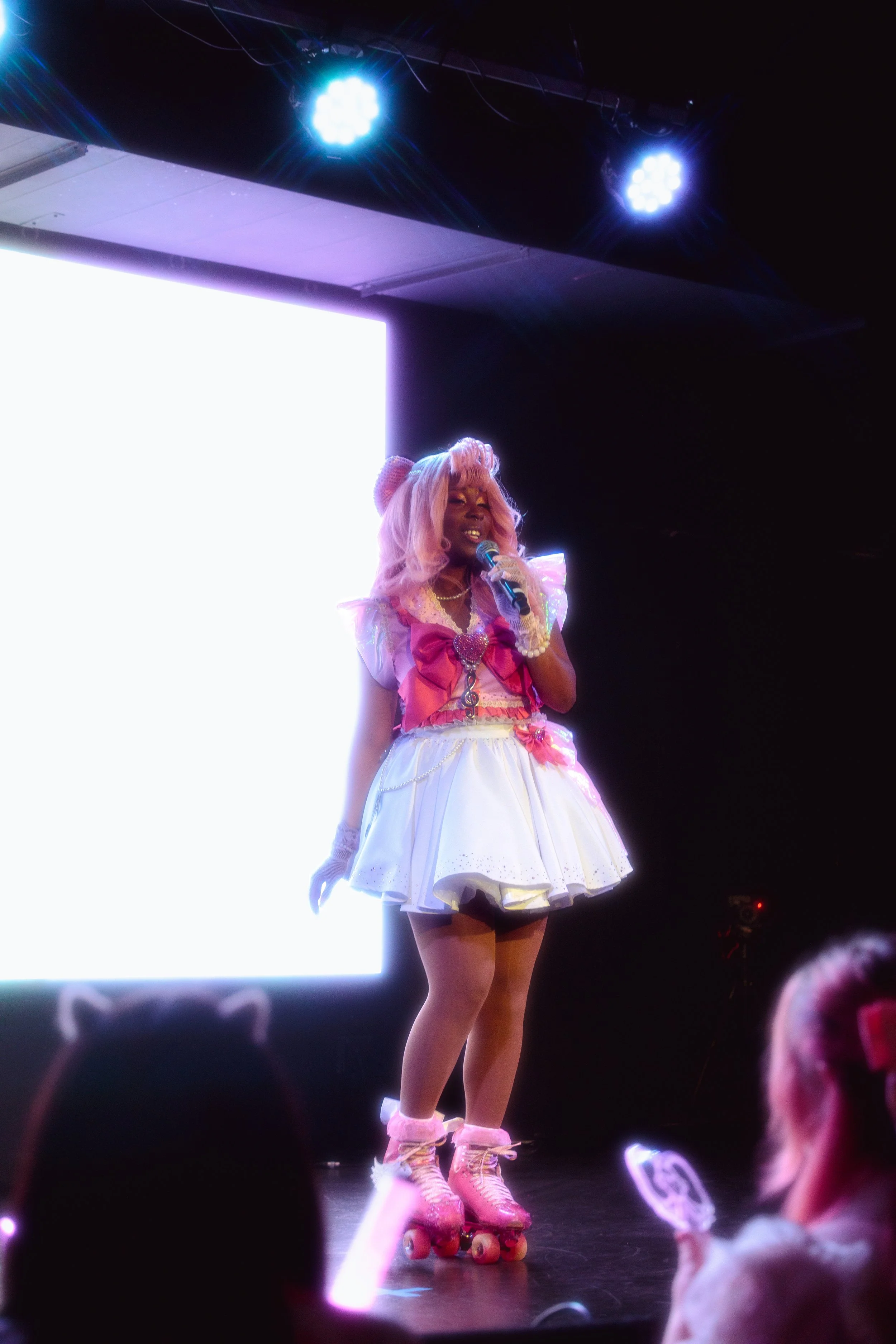 A performer dressed in a pink and white costume with roller skates on stage under blue stage lights, holding a microphone, backlit by a bright white screen.