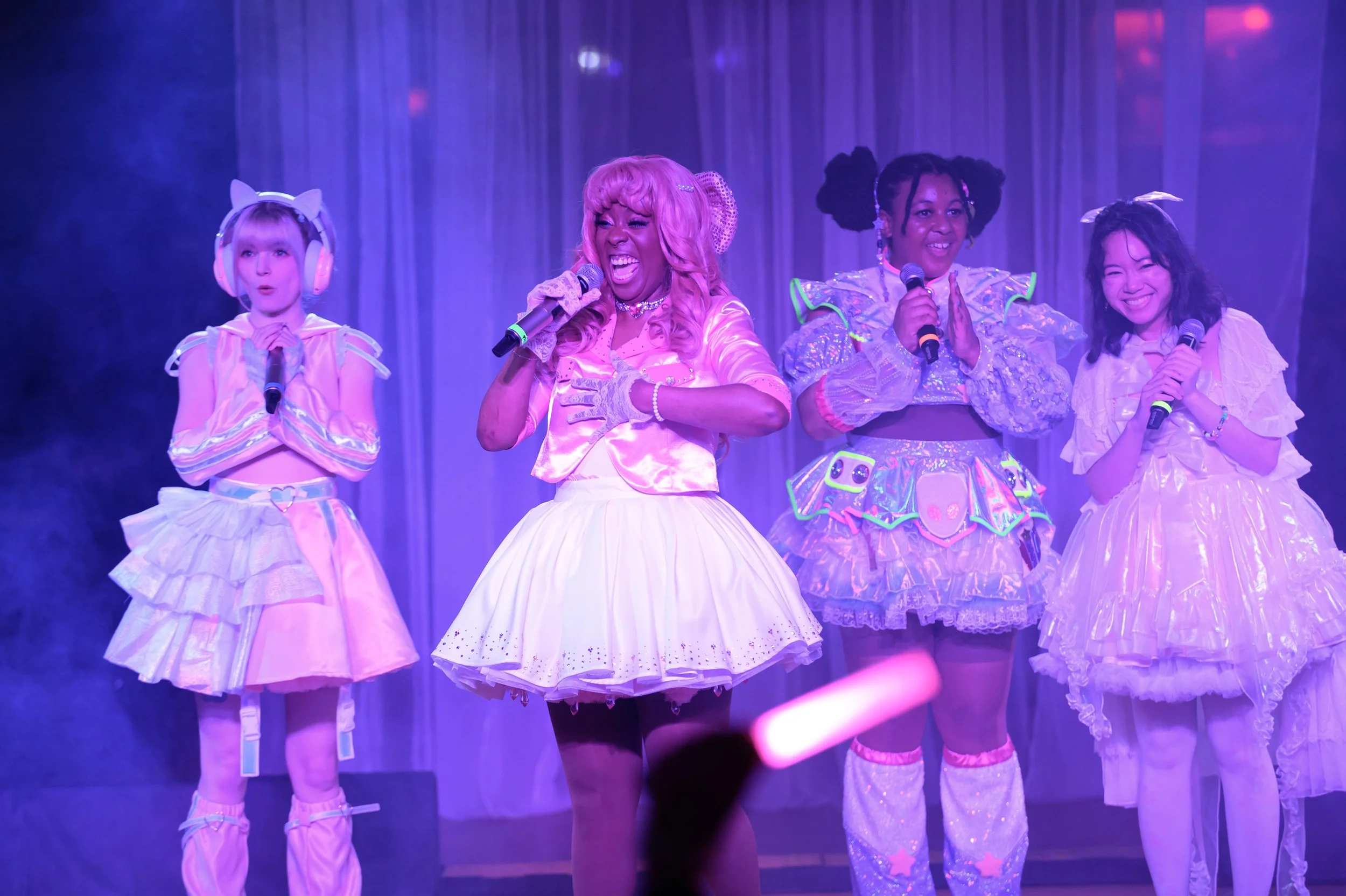 Four performers on stage dressed in pastel-colored, fantasy-inspired costumes, holding microphones, with purple stage lighting and a curtain backdrop.
