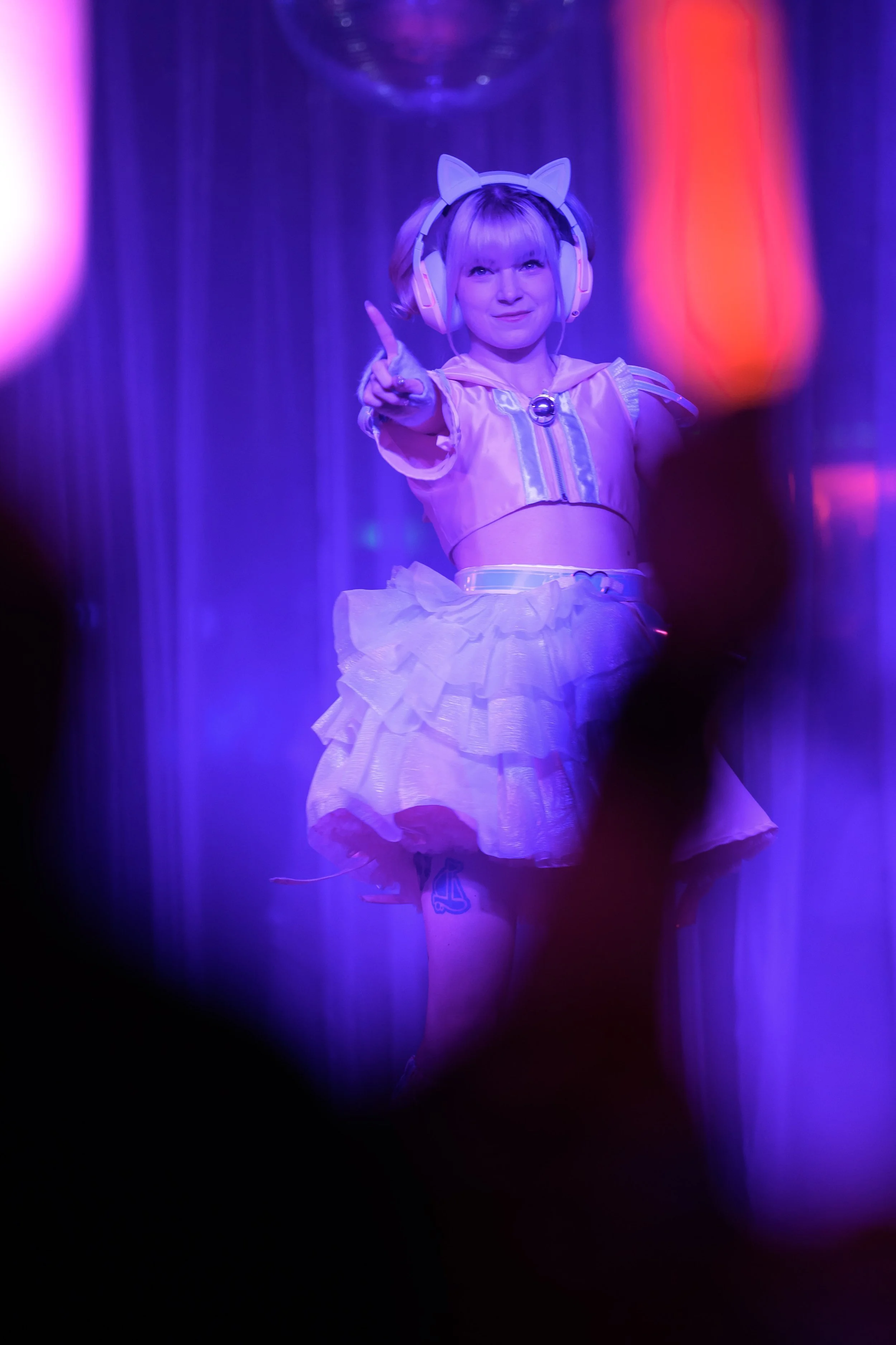 Young girl with cat ear headphones and pink top, performing on stage with purple lighting, pointing upwards.