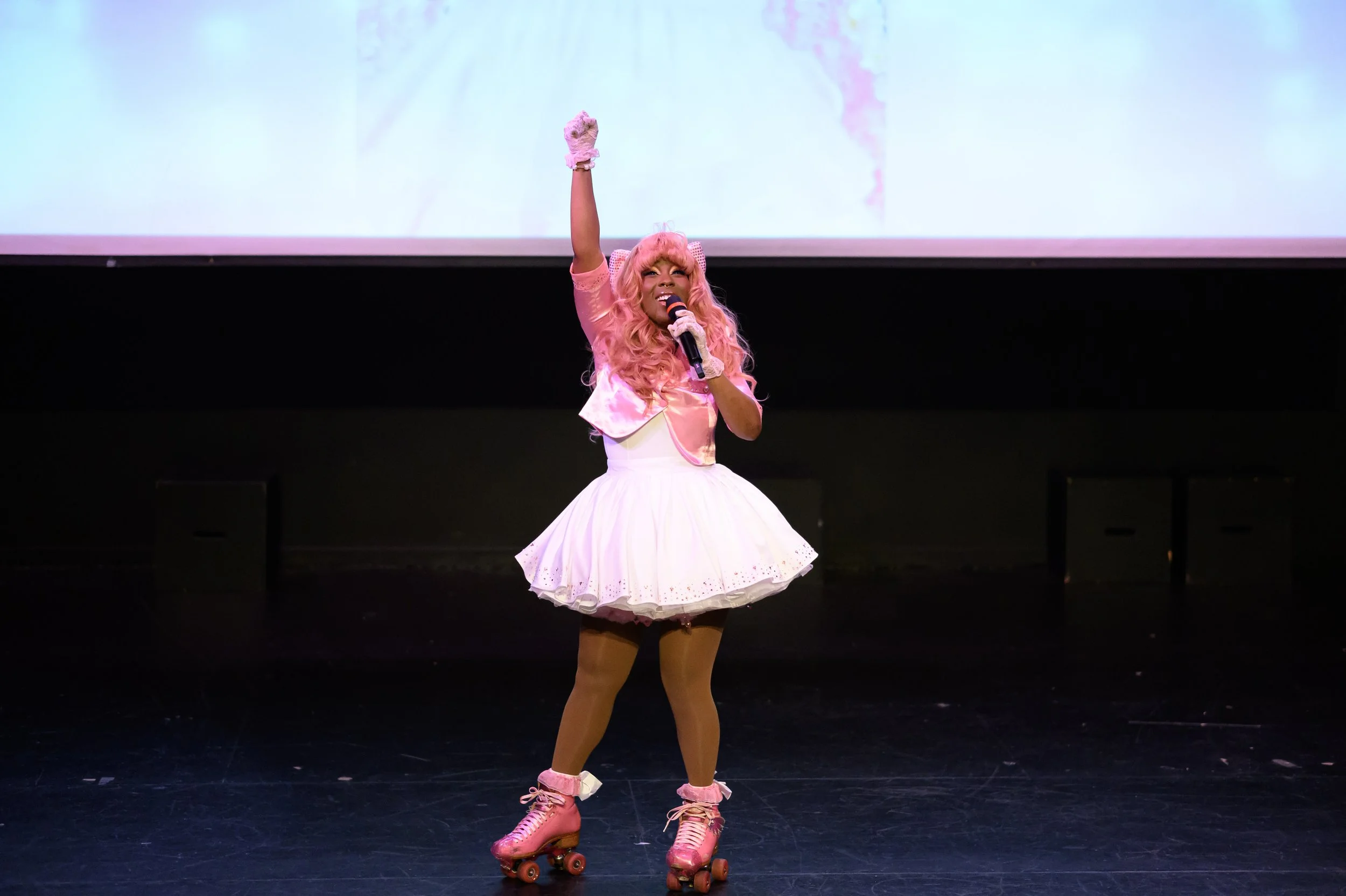 Person in pink costume on roller skates performing on stage with a microphone.