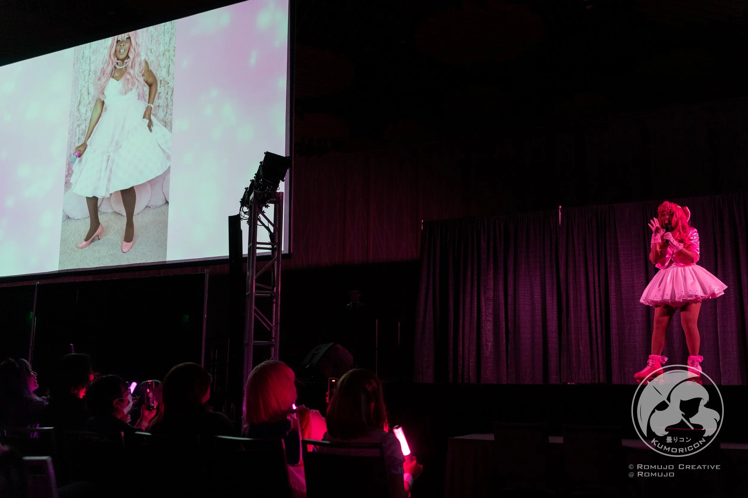 A person dressed in a pink costume with a puffy skirt, standing on stage under pink lighting, speaking to an audience. A large screen behind shows a photograph of a woman in a white dress with pink hair, pink high heels, and jewelry.