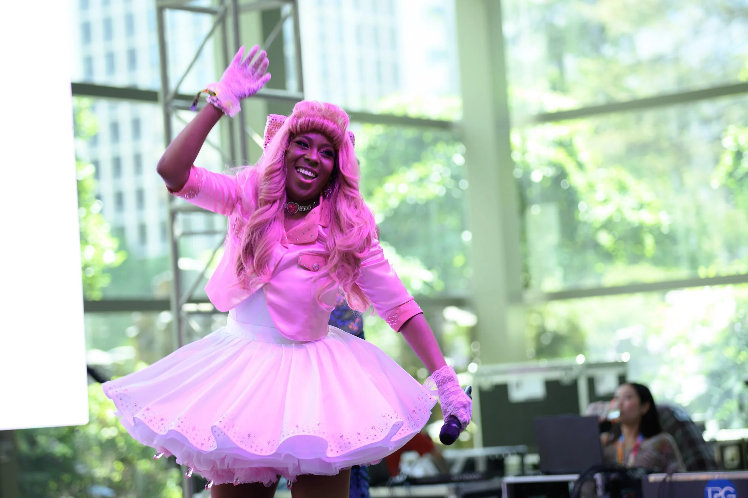 A woman with long, wavy pink hair wearing a pink dress and gloves, smiling, waving, and holding a microphone on stage with green trees and a woman with a laptop in the background.