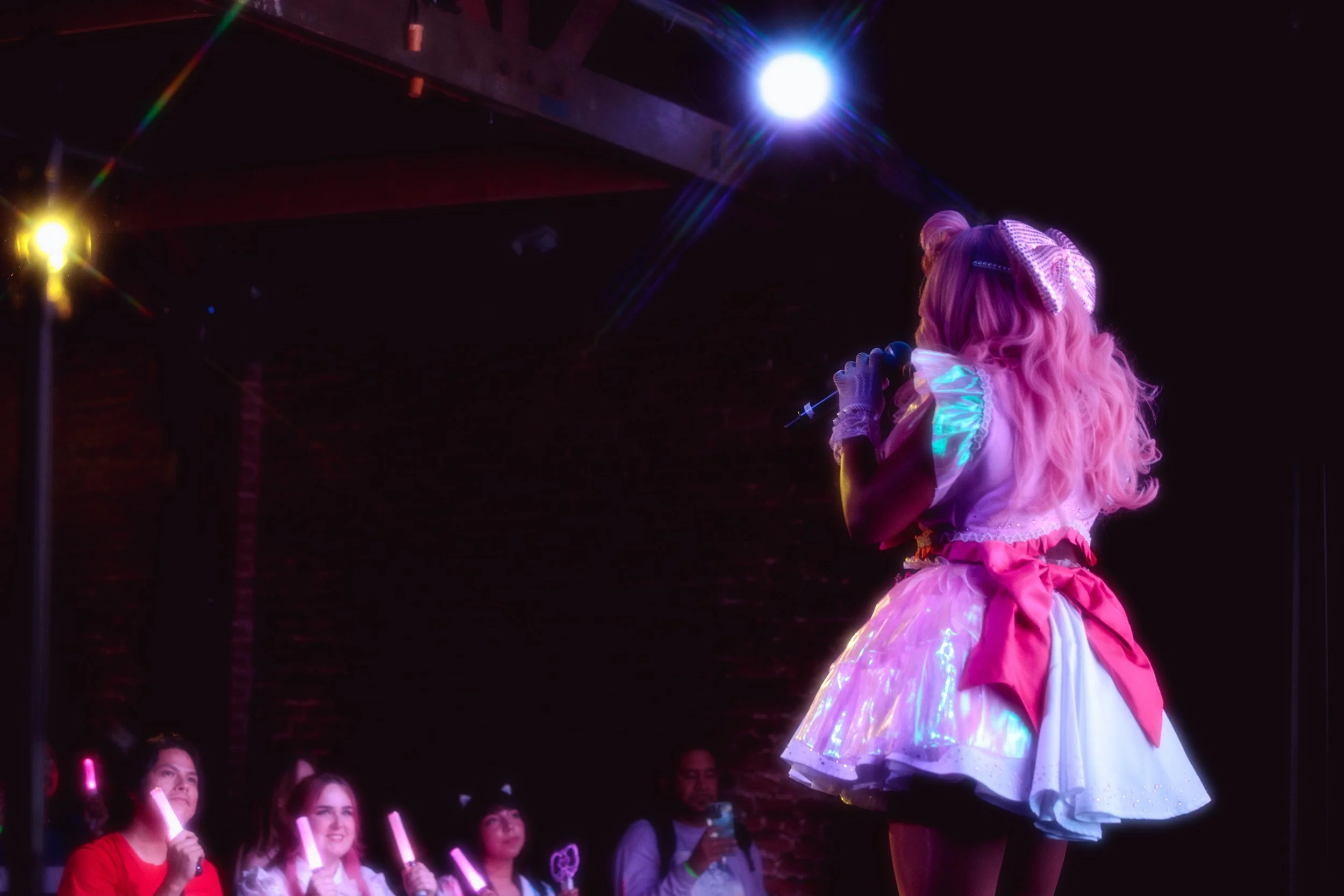 A girl with pink hair in a princess costume performing on stage with a microphone. Audience members in the front are holding glow sticks and watching her.
