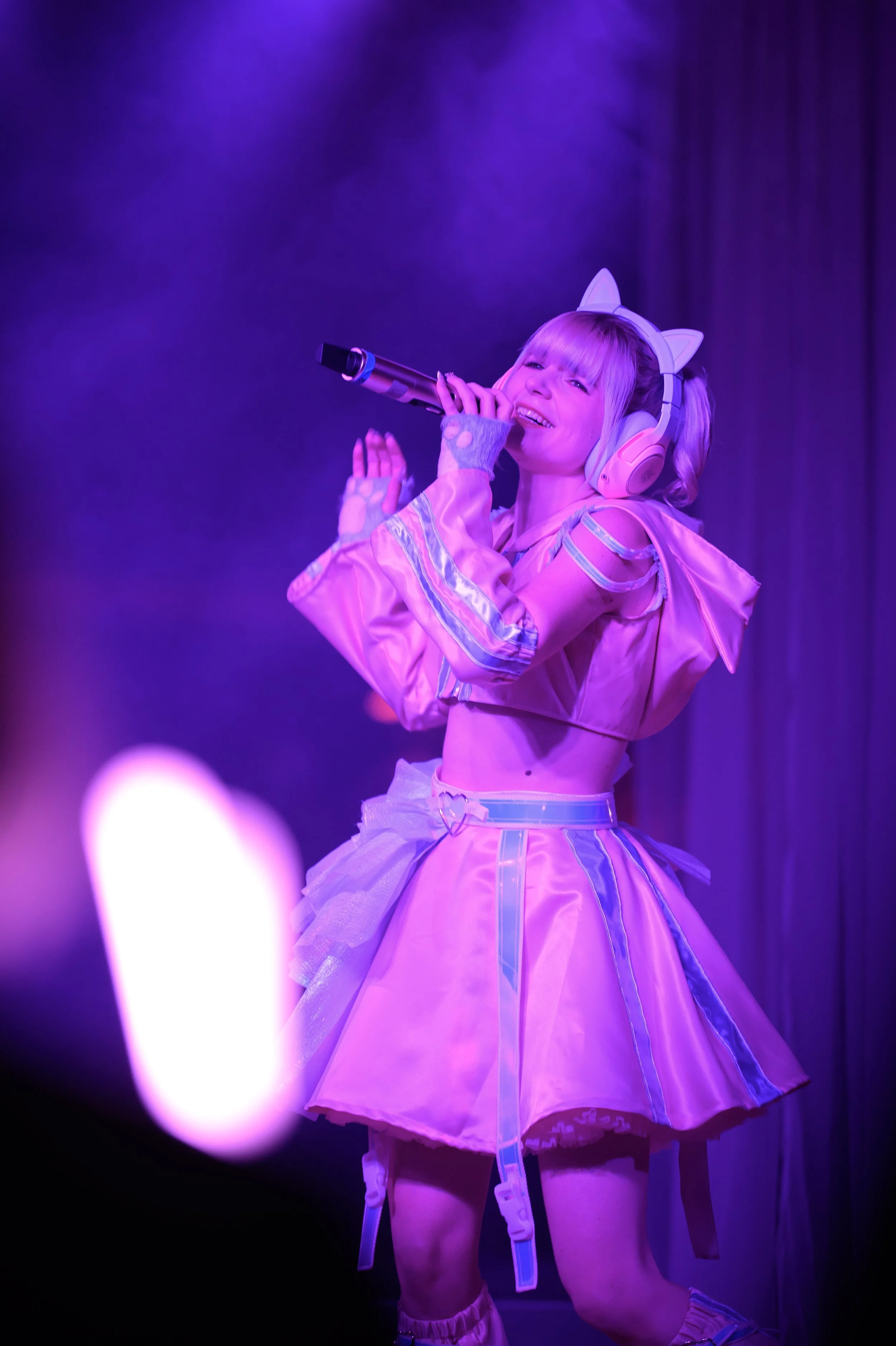 Performer on stage wearing a pink costume with cat-ear headphones singing into a microphone under purple stage lighting.