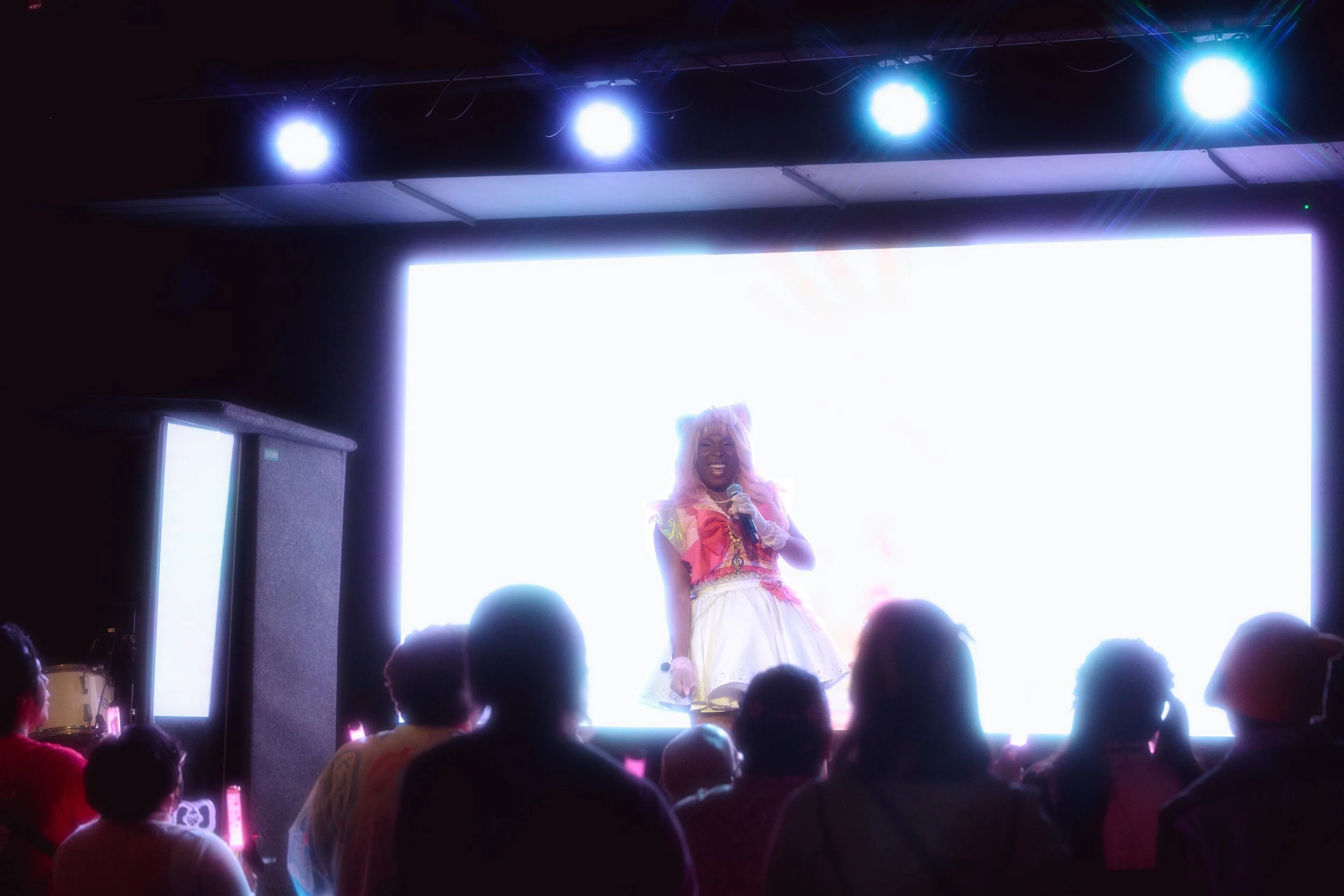 Performer on stage with a bright digital screen behind, holding a microphone, wearing a colorful costume with a skirt, and smiling, while an audience watches.