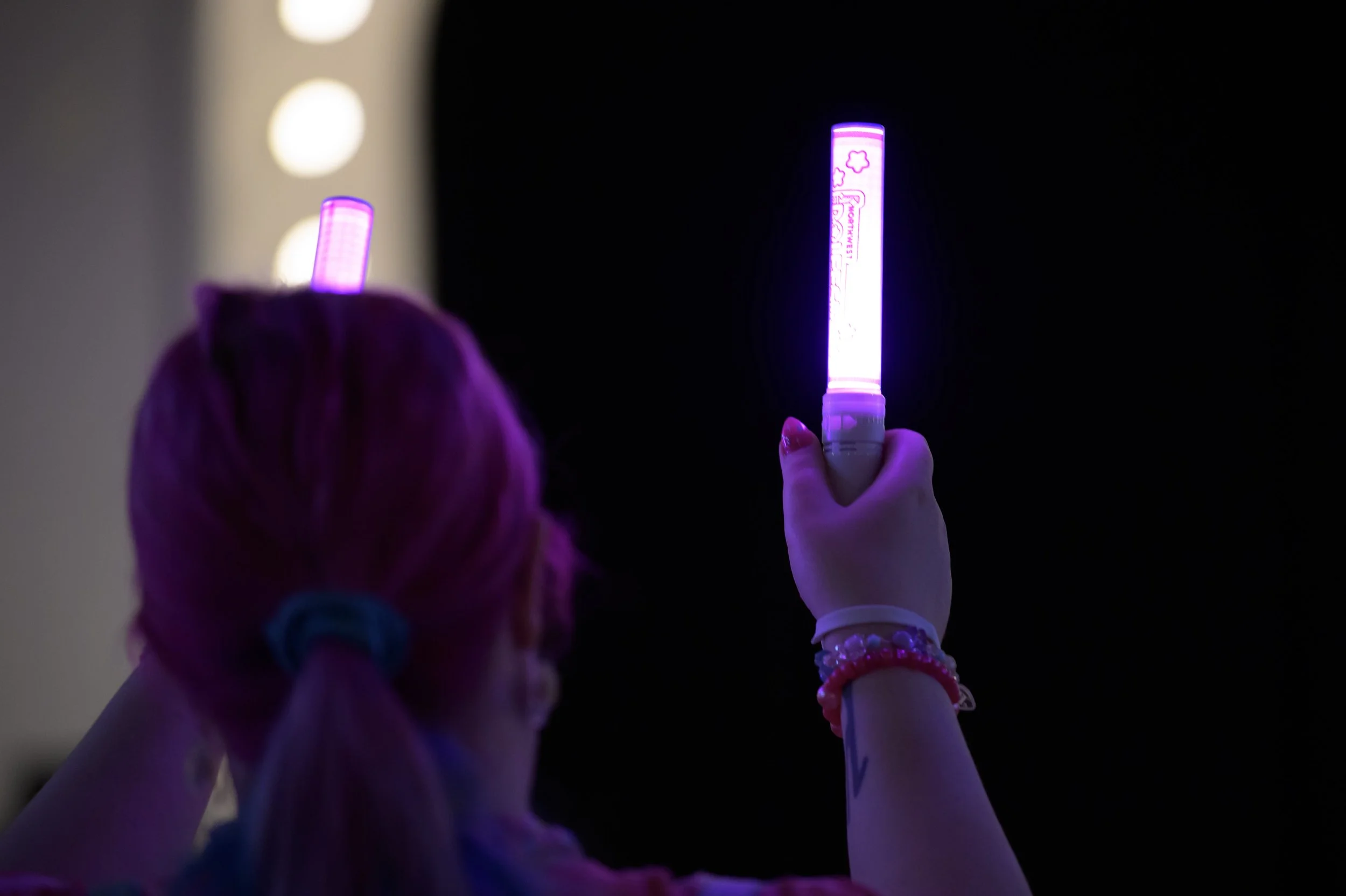 Person holding a pink light-up glow stick in a dark environment, with a person with pink hair and a hair tie visible, and blurred circular lights in the background.