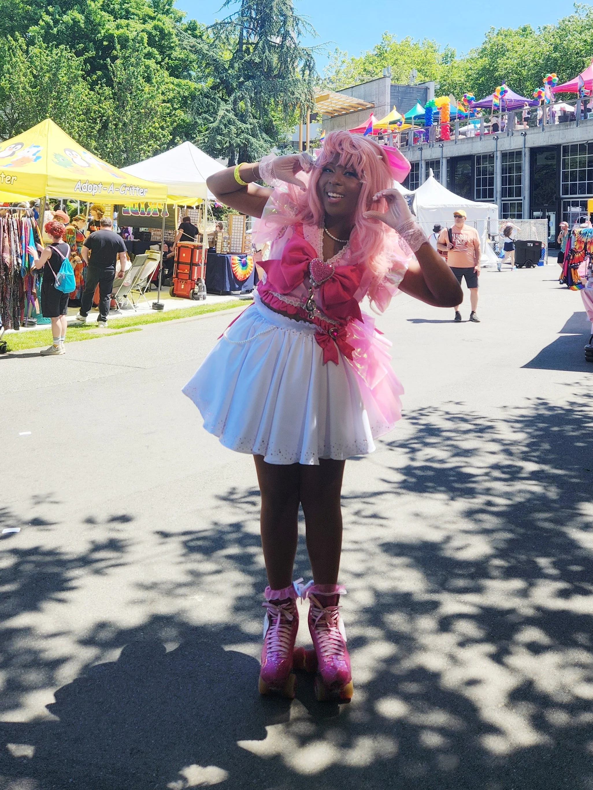 Person in pink and white costume skating at outdoor festival with vendors, tents, and trees in the background.
