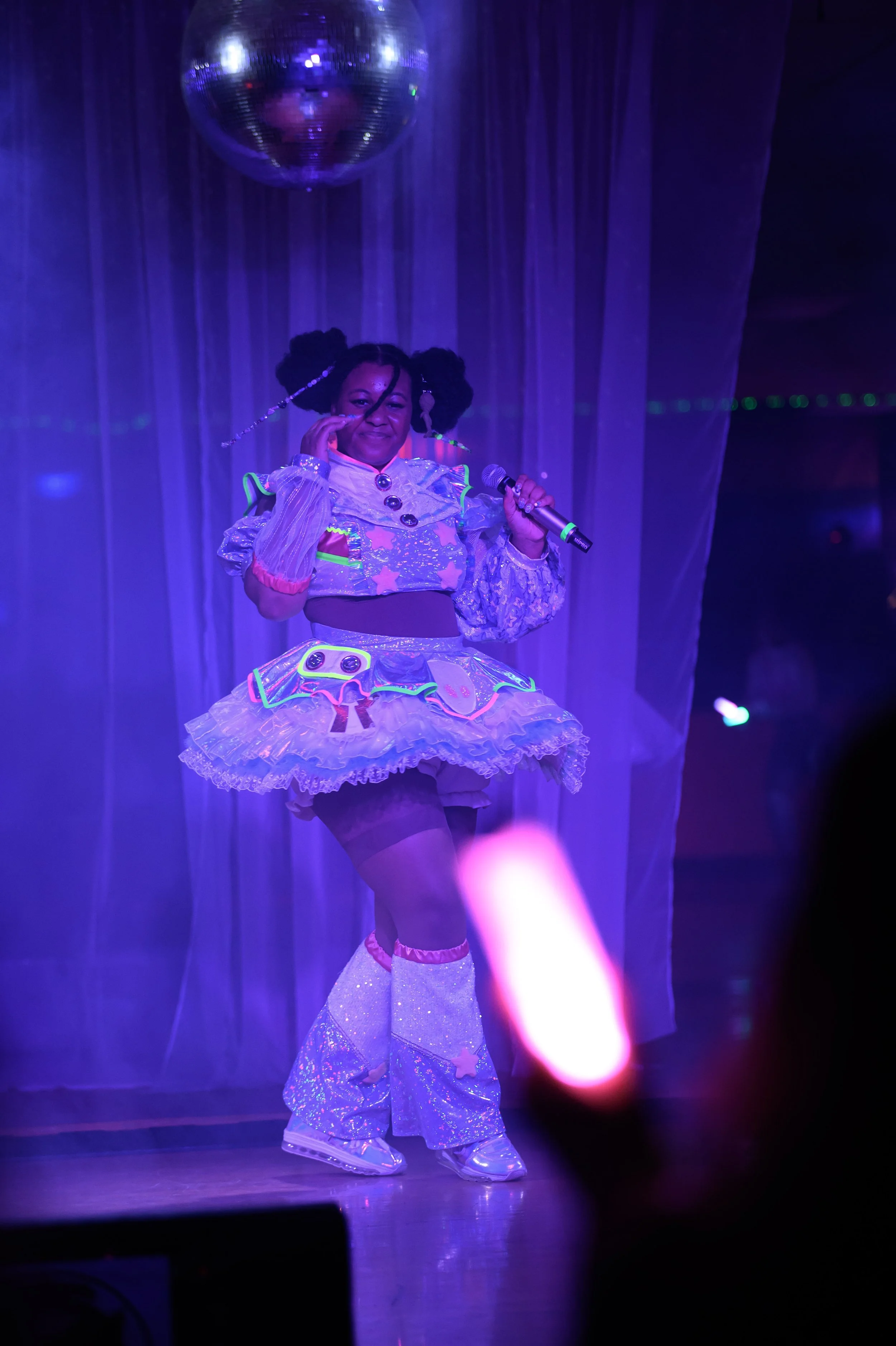 A woman dressed in a colorful, shiny costume with a holographic skirt, oversized boots, and a glittery top, performing on stage with a microphone, under a disco ball with purple lighting.