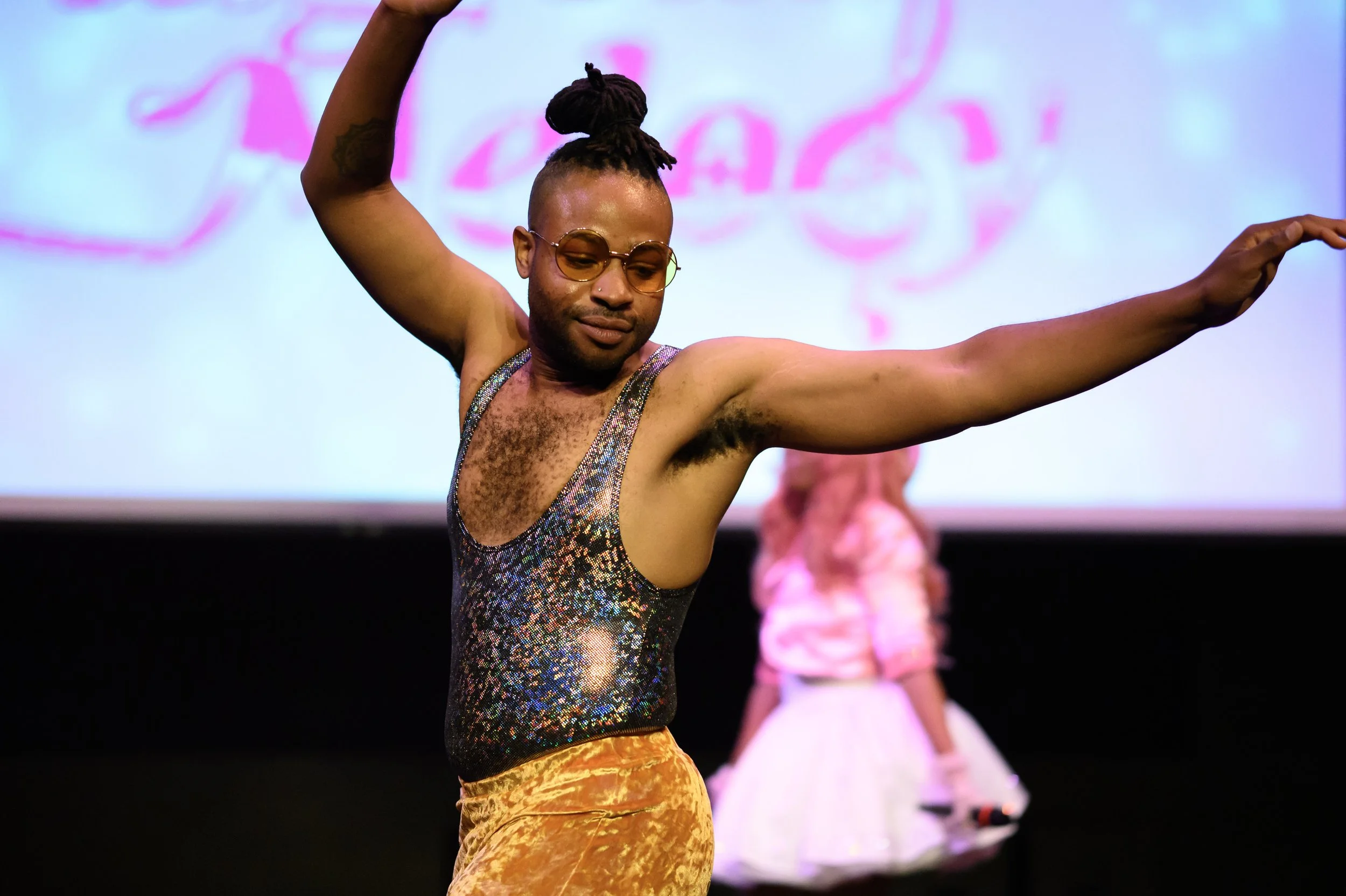 A person with dark hair styled in braids and glasses, dancing on stage. They are wearing a sleeveless, glittery top and yellow velvet pants. In the background, another person in a pink jacket and white skirt is holding a microphone.