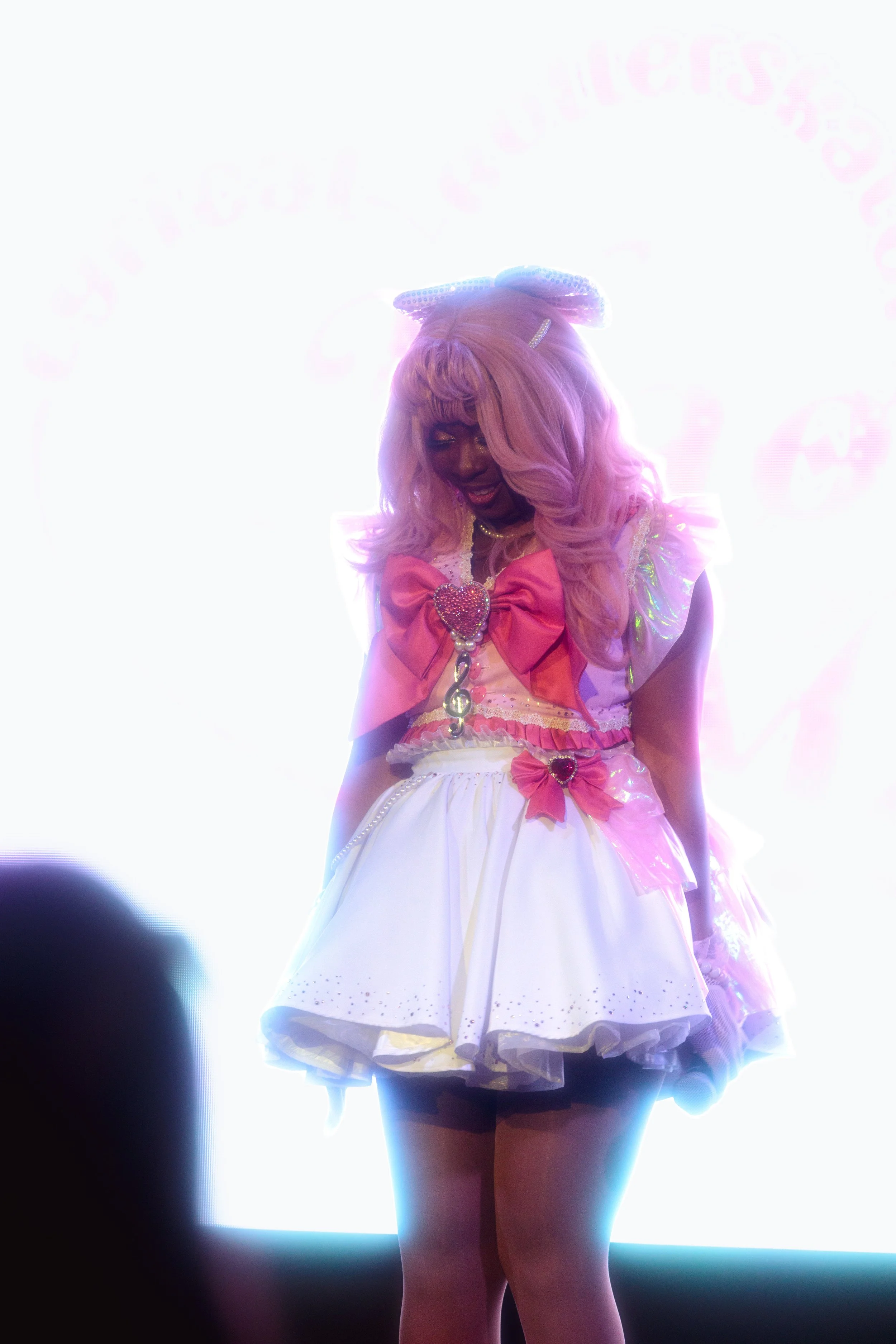 Person dressed as a magical girl, with pink hair, wearing a pink and white costume with large pink bows, a heart-shaped accessory, and holding a microphone, on a brightly lit stage.