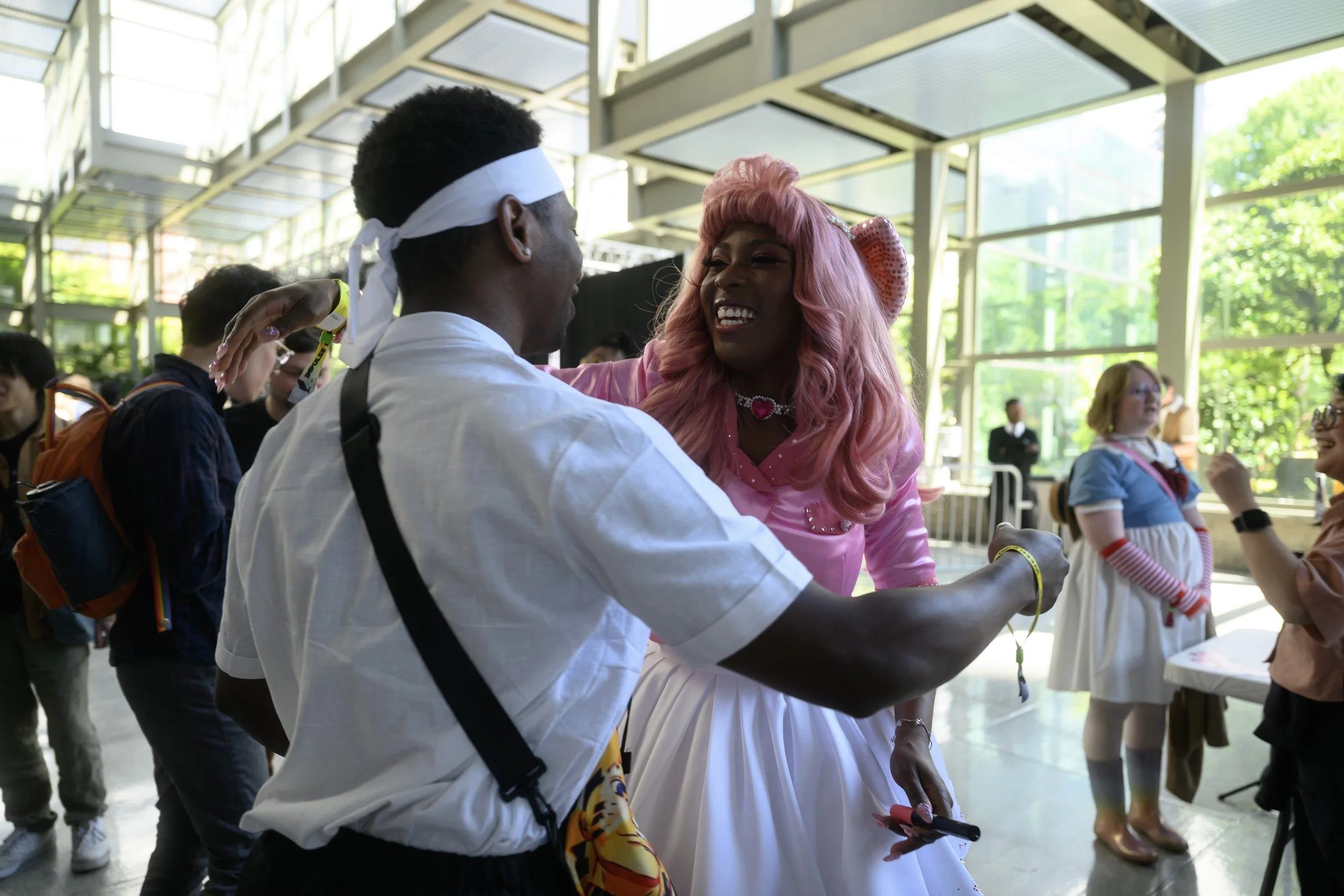 Two people talking in a large glass-walled lobby, with others in cosplay costumes around them.